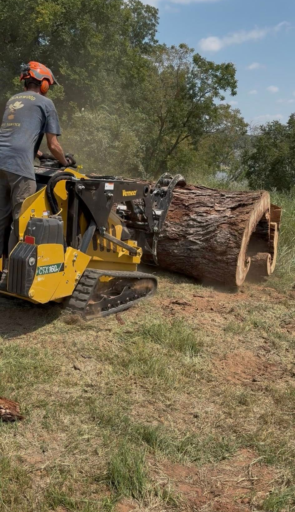 A person operating a yellow skid steer, moving a large log in an outdoor setting.