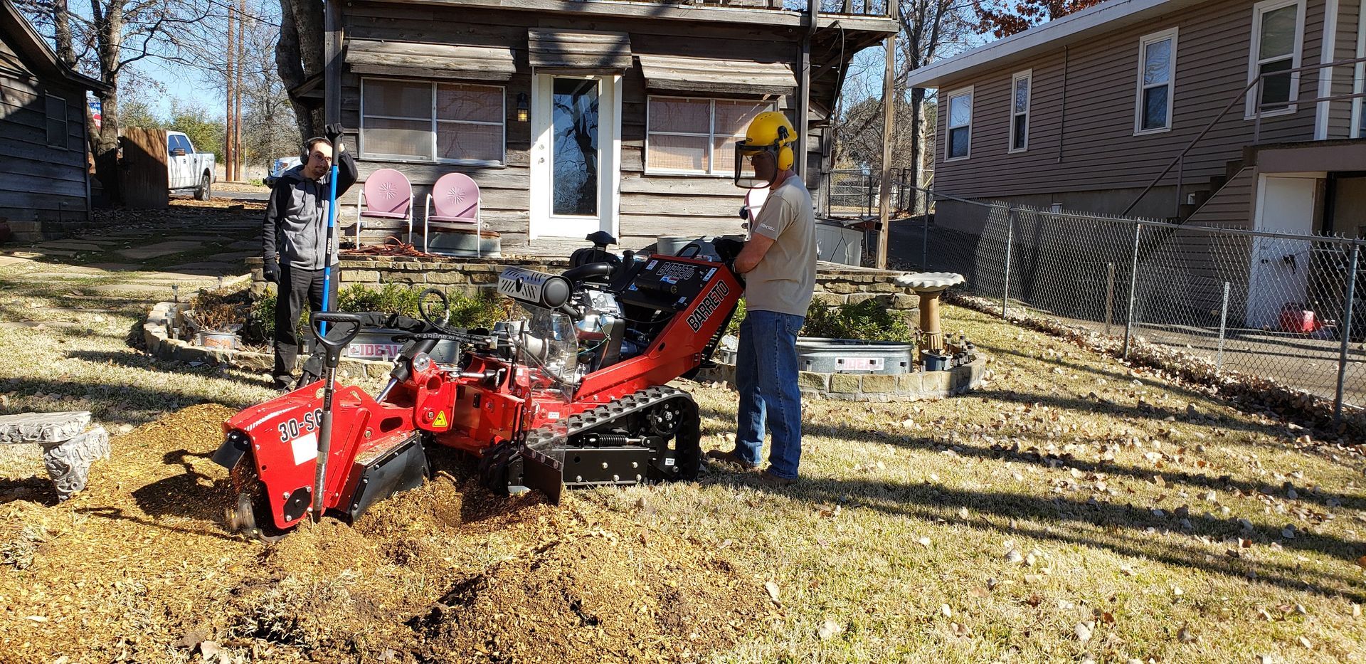 Two people using a red stump grinder in a yard. One wears ear protection. A house is in the background.