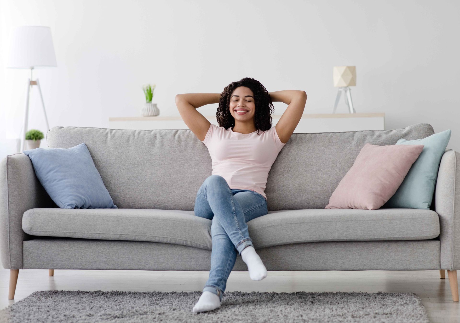 Person relaxing on a gray sofa in a bright living room, smiling with hands behind head.