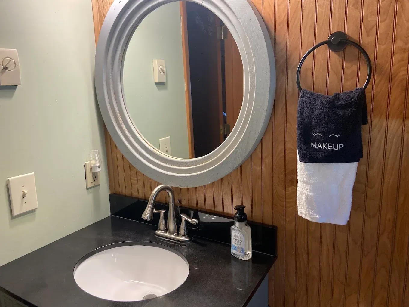 Bathroom vanity with round mirror, white sink, black countertop, wood wall, and hanging towel organizer