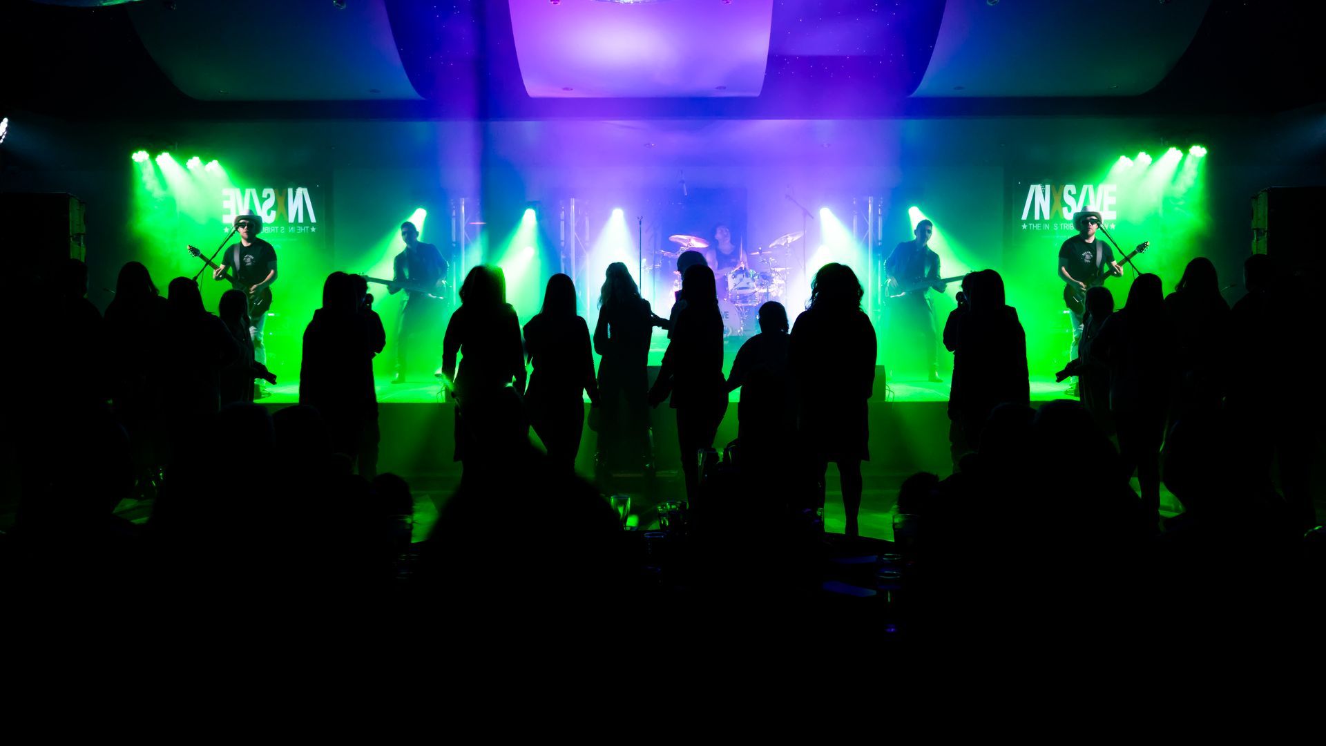 Band performing on stage, silhouetted against green and purple lights. Audience in front.
