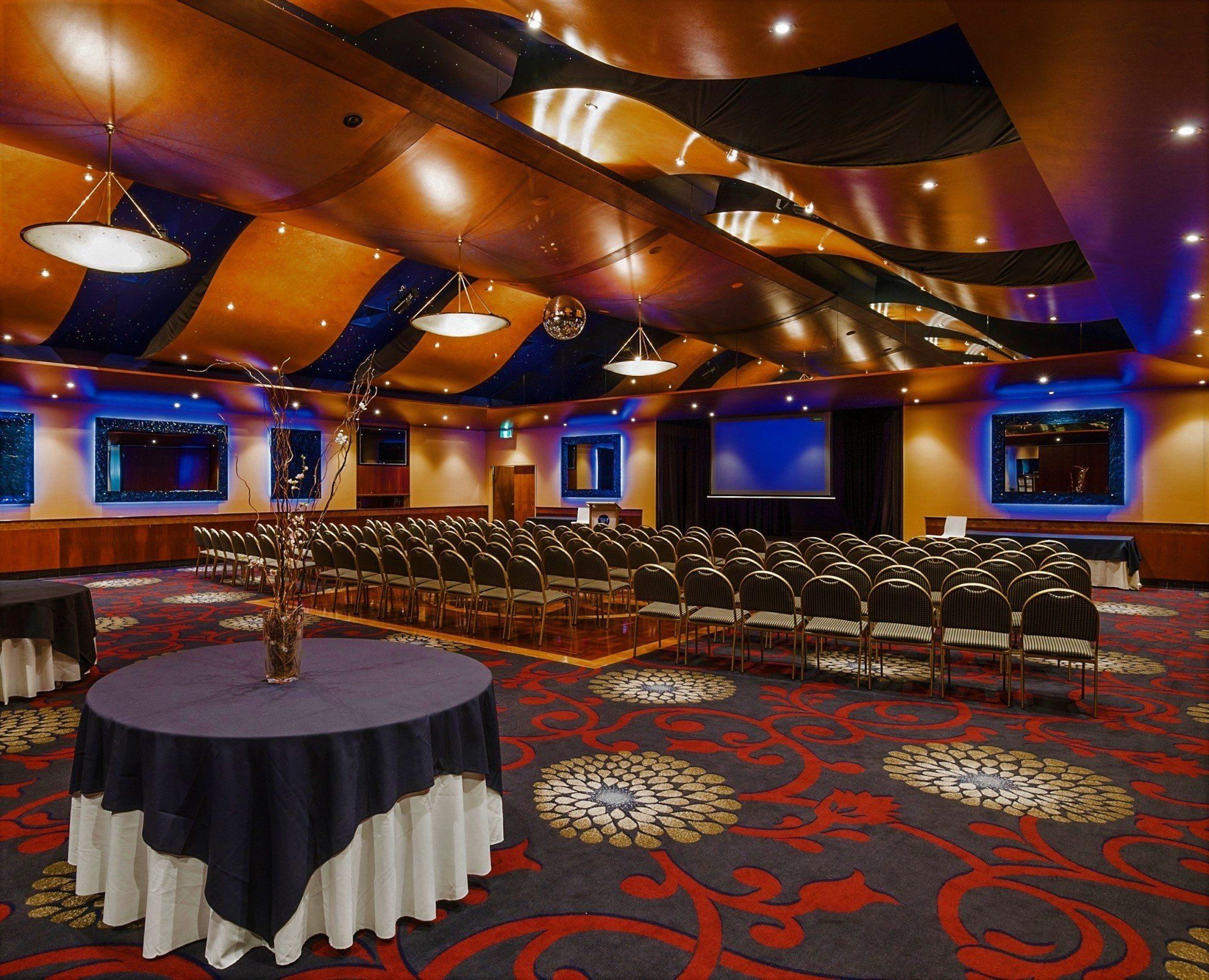 Auditorium with rows of chairs, round tables, patterned carpet, and wavy ceiling decorations.