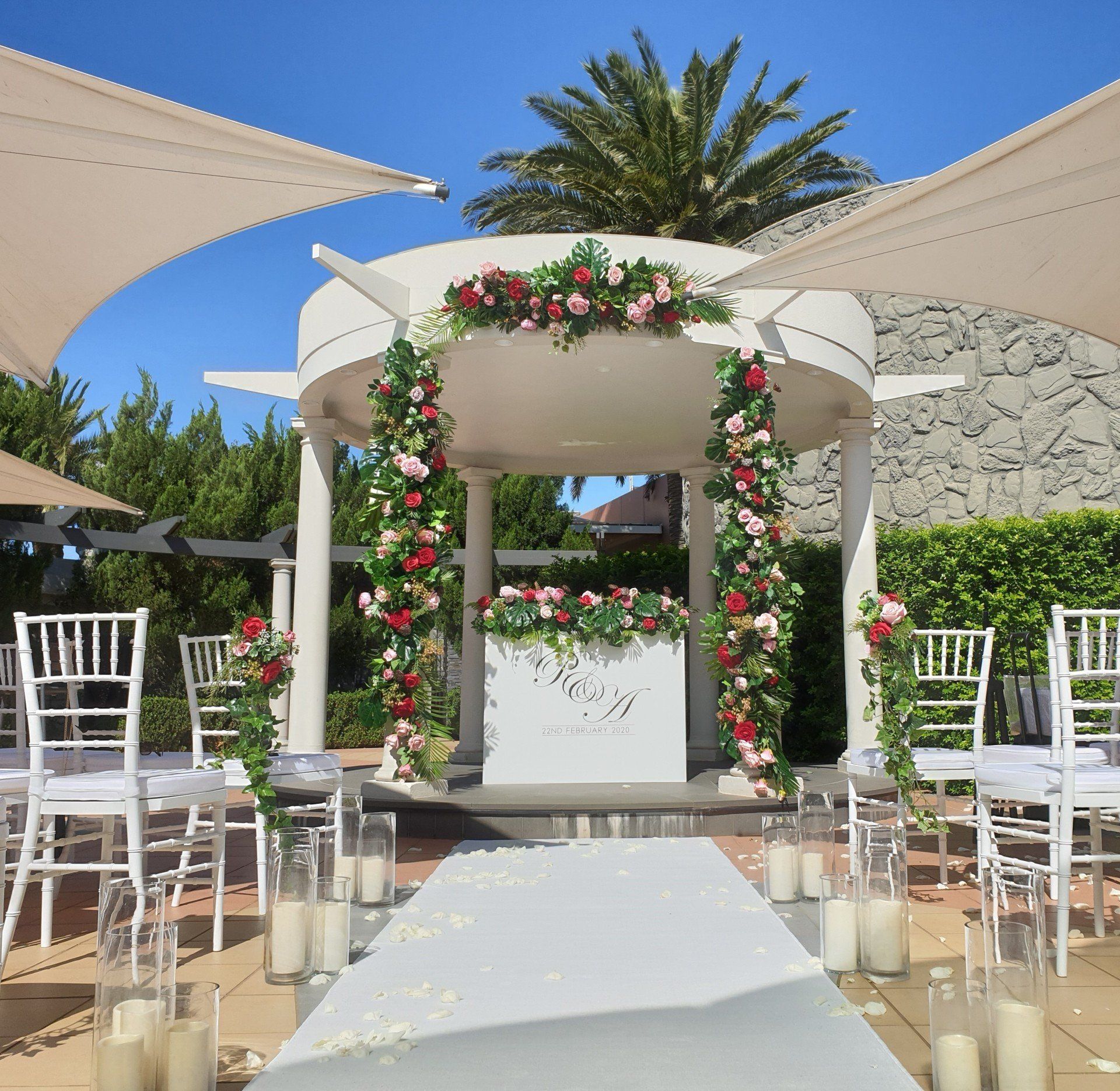 Wedding ceremony setup: White chairs, floral arch, aisle runner, and candles on a sunny day.