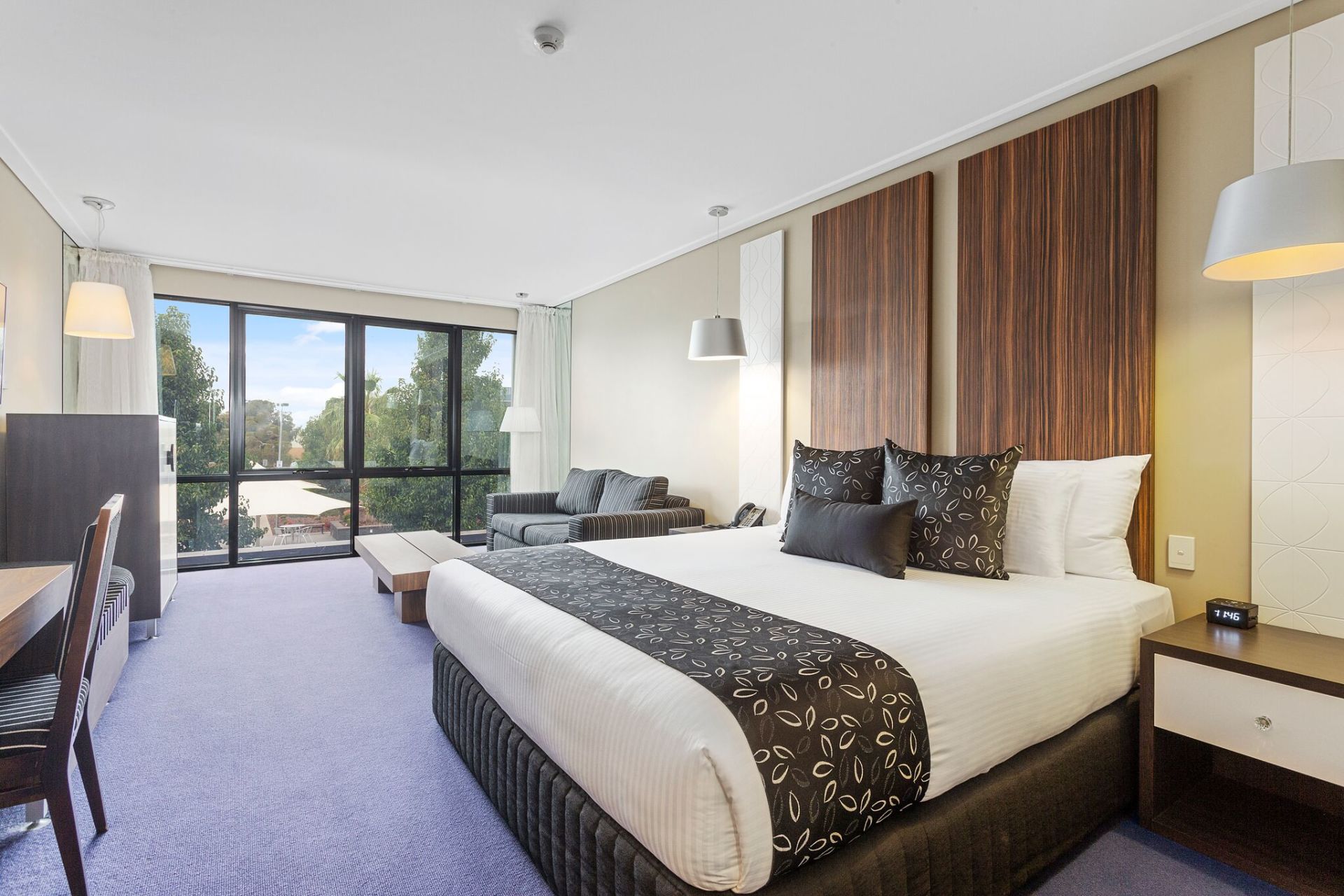 Hotel bedroom with king-size bed, sofa, desk, and large window with outside view; neutral colors with accent wall.