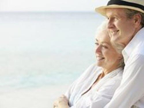 Elderly couple embracing on a beach, both smiling, overlooking the ocean.