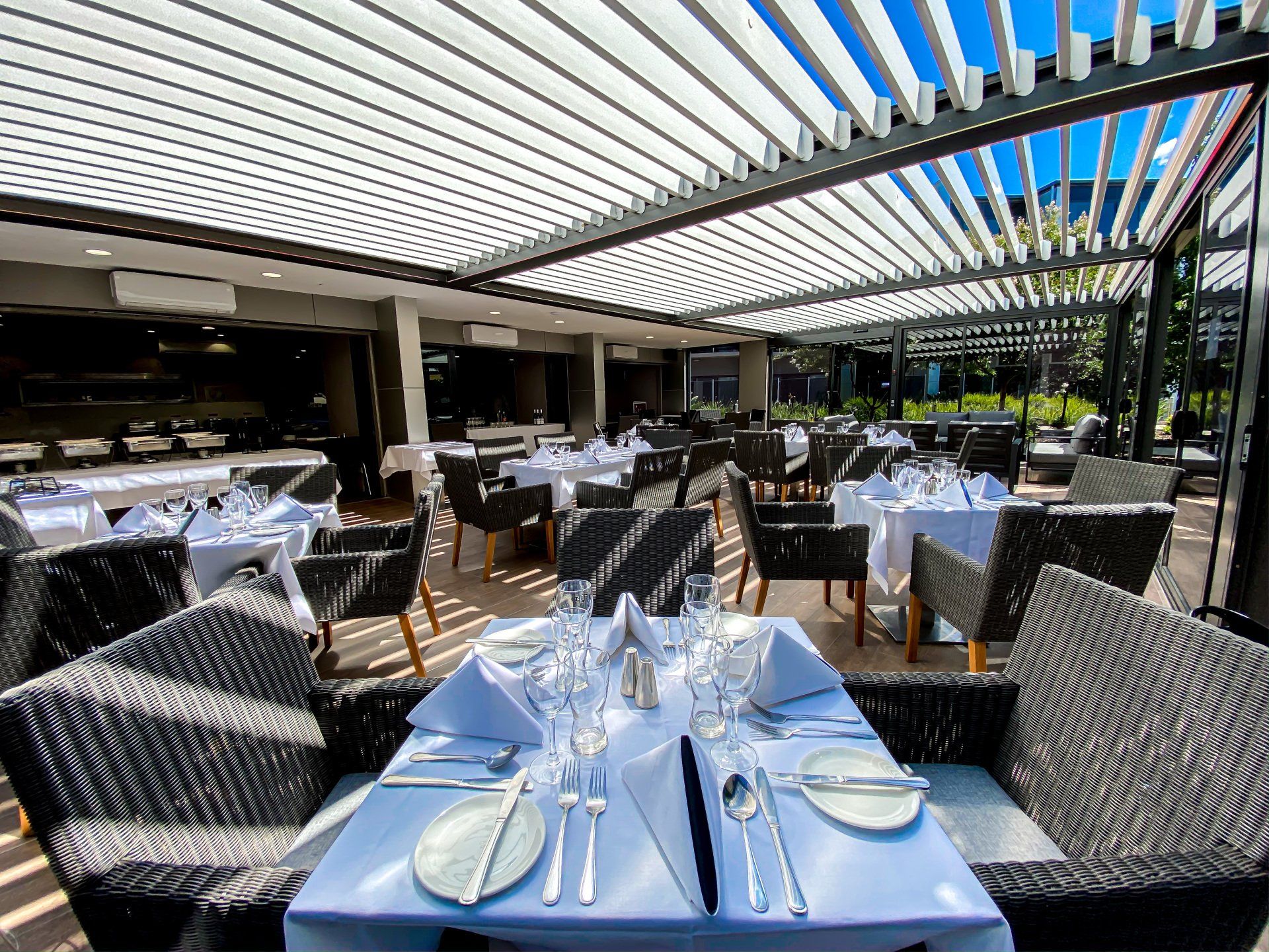 Restaurant with white tablecloths, dark chairs, under a slatted pergola.