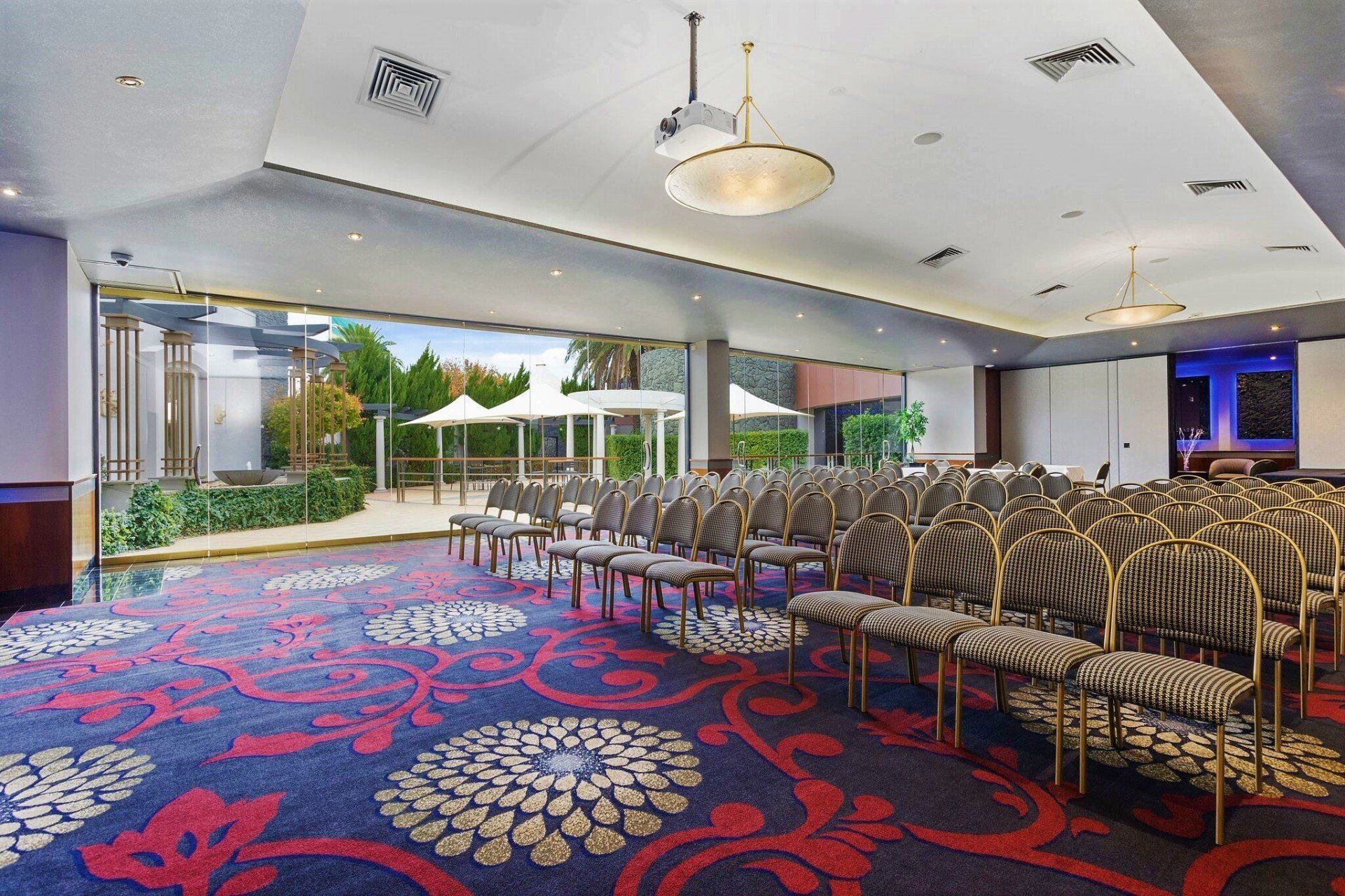 A conference room with rows of chairs, a large floral carpet, and a view of an outdoor area through sliding glass doors.