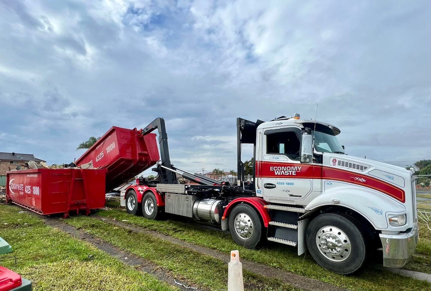 A Large Truck With A Red Dumpster Attached To It Is Parked In A Grassy Field — Economy Waste Group | Skip Bins Central Coast In Bateau Bay, NSW