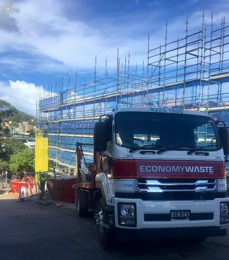 A Red And White Truck Is Parked In Front Of A Building With Scaffolding — Economy Waste Group | Skip Bins Central Coast In The Entrance, NSW