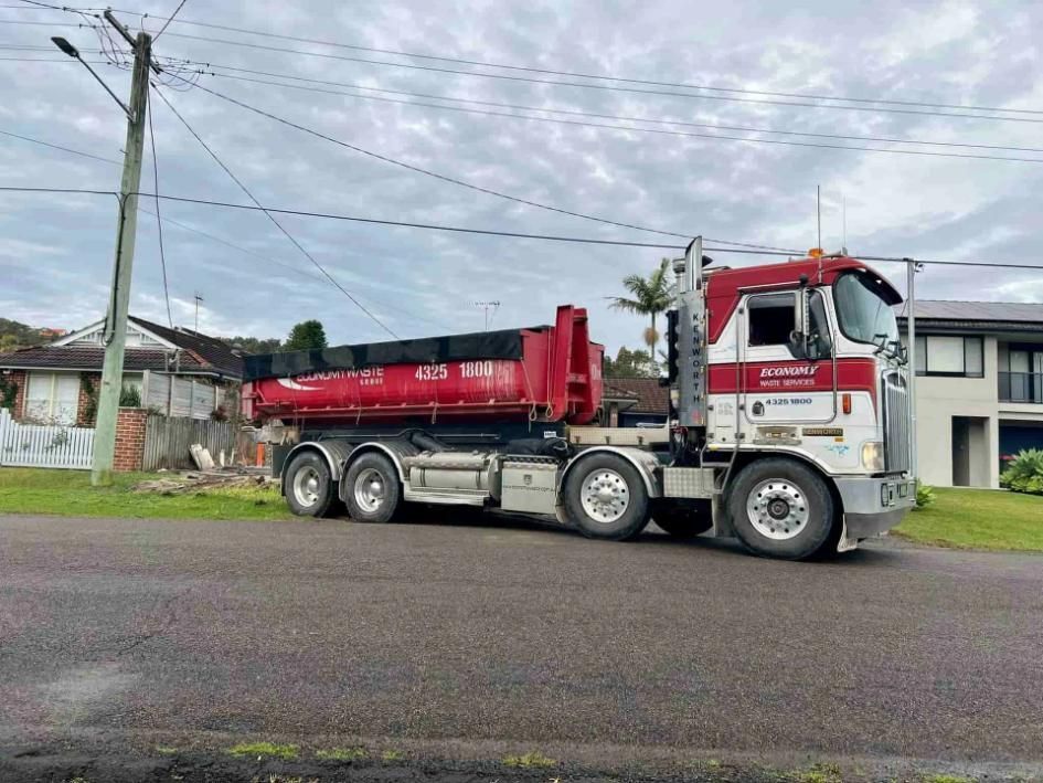 A Red Dump Truck Is Driving Down A Street Next To A House — Economy Waste Group | Skip Bins Central Coast In Terrigal, NSW