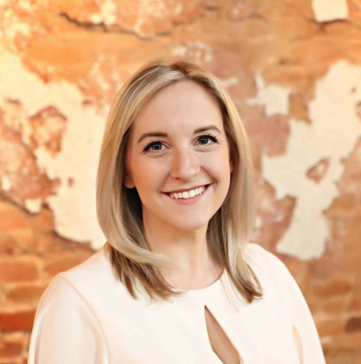 A woman in a white shirt is smiling in front of a brick wall.
