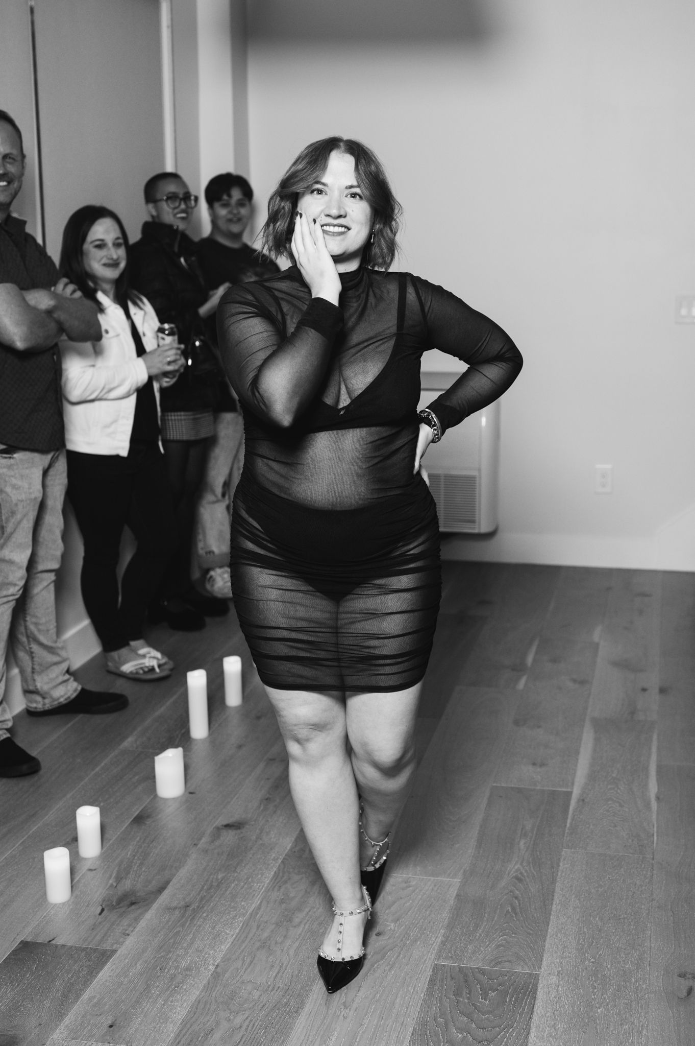 A woman wearing a sheer dress stands confidently in front of an audience, exuding elegance and poise.