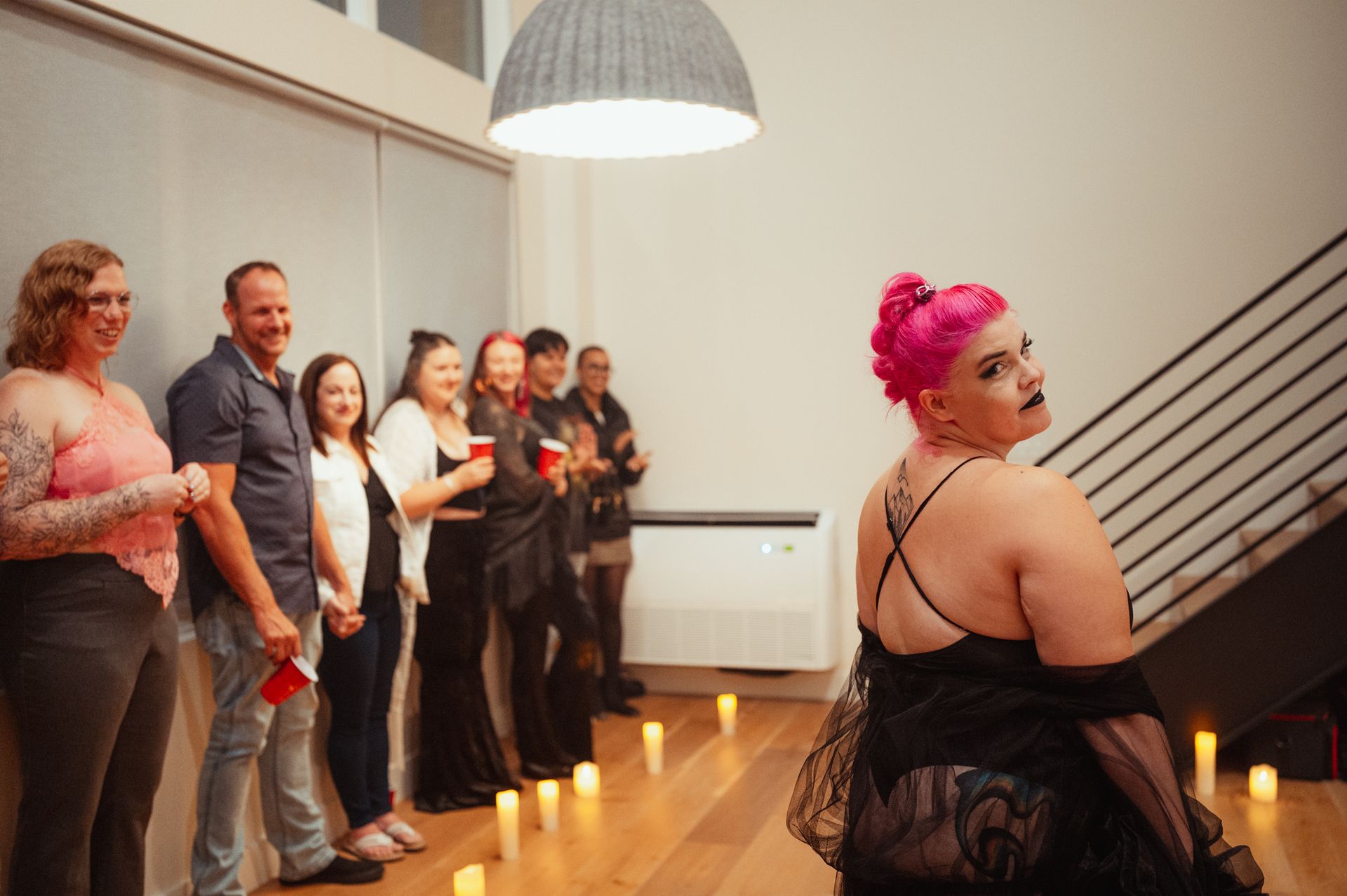 A woman with vibrant pink hair, dressed in black, stands in front of a crowd, exuding charisma and presence.