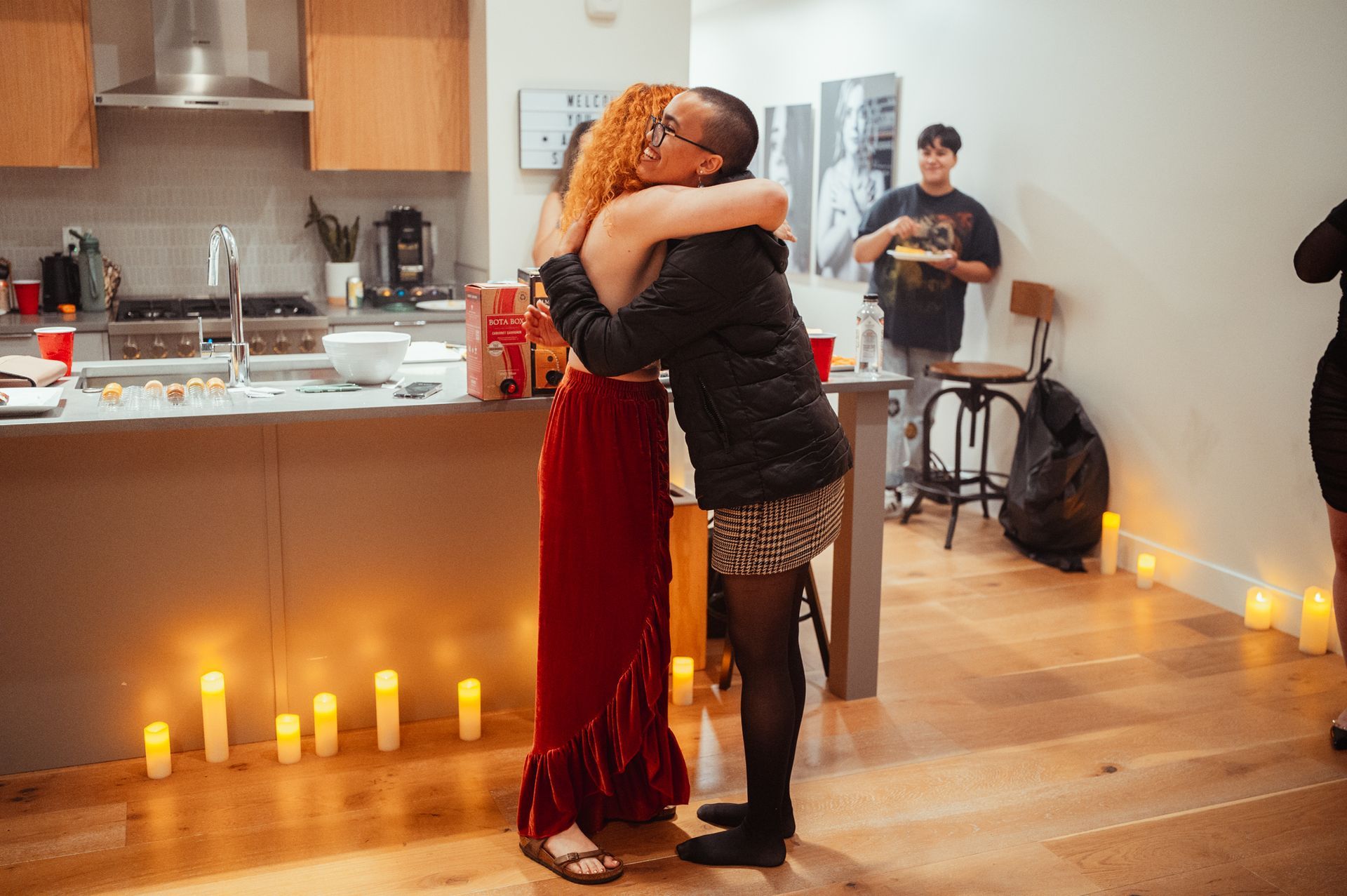 Two women embrace warmly in a cozy kitchen, illuminated by the soft glow of candles, creating a serene atmosphere.