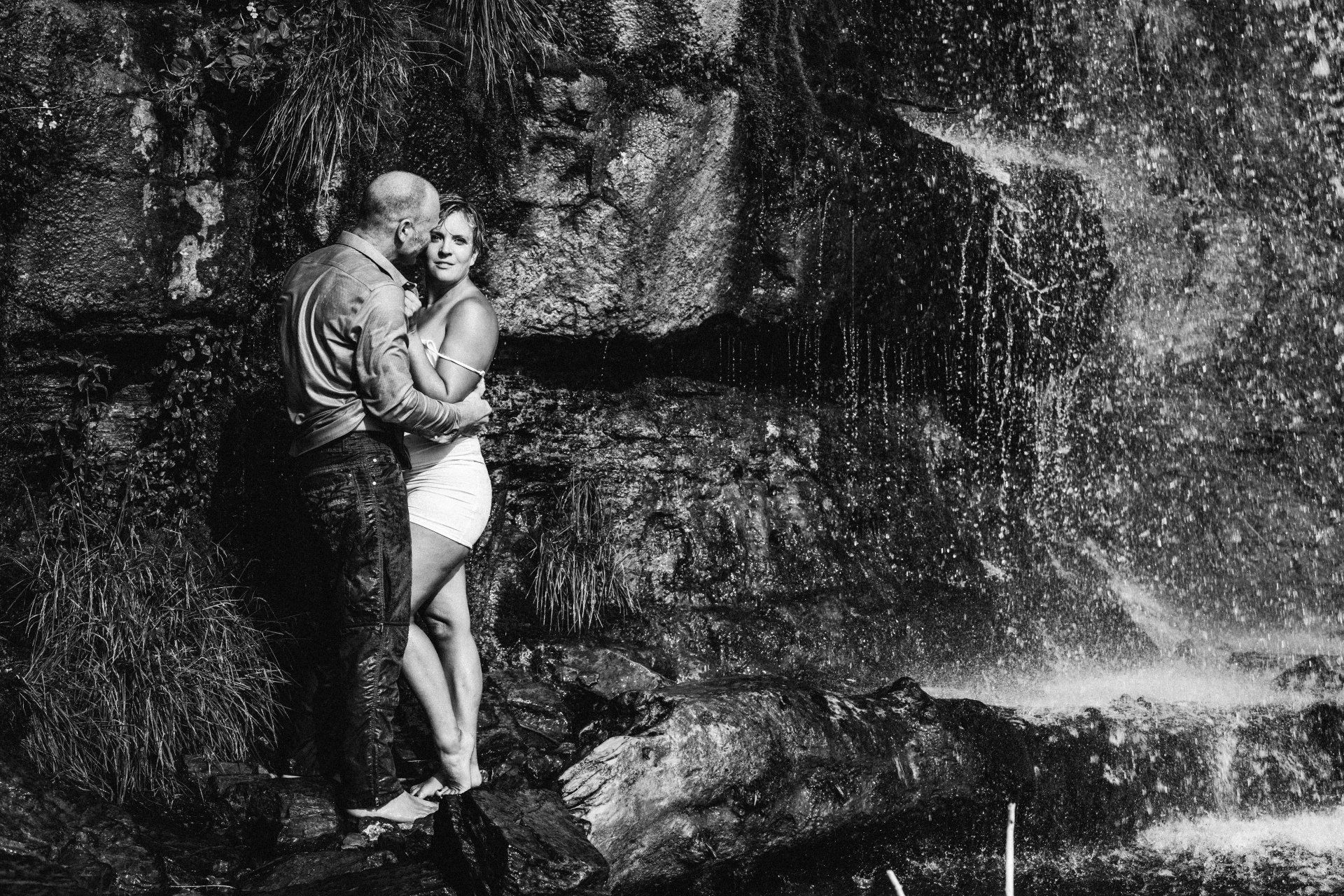 A couple standing in front of a waterfall, enjoying the scenic beauty and feeling the mist on their faces.