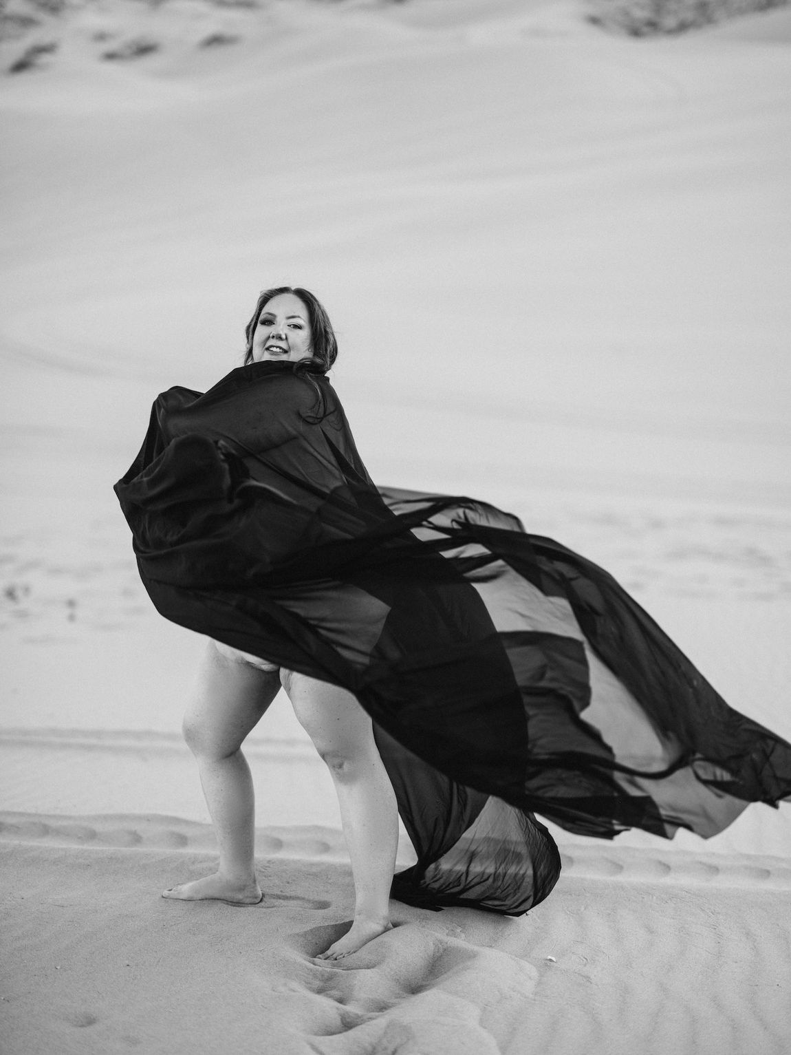A woman in a black dress stands gracefully on the sandy beach.