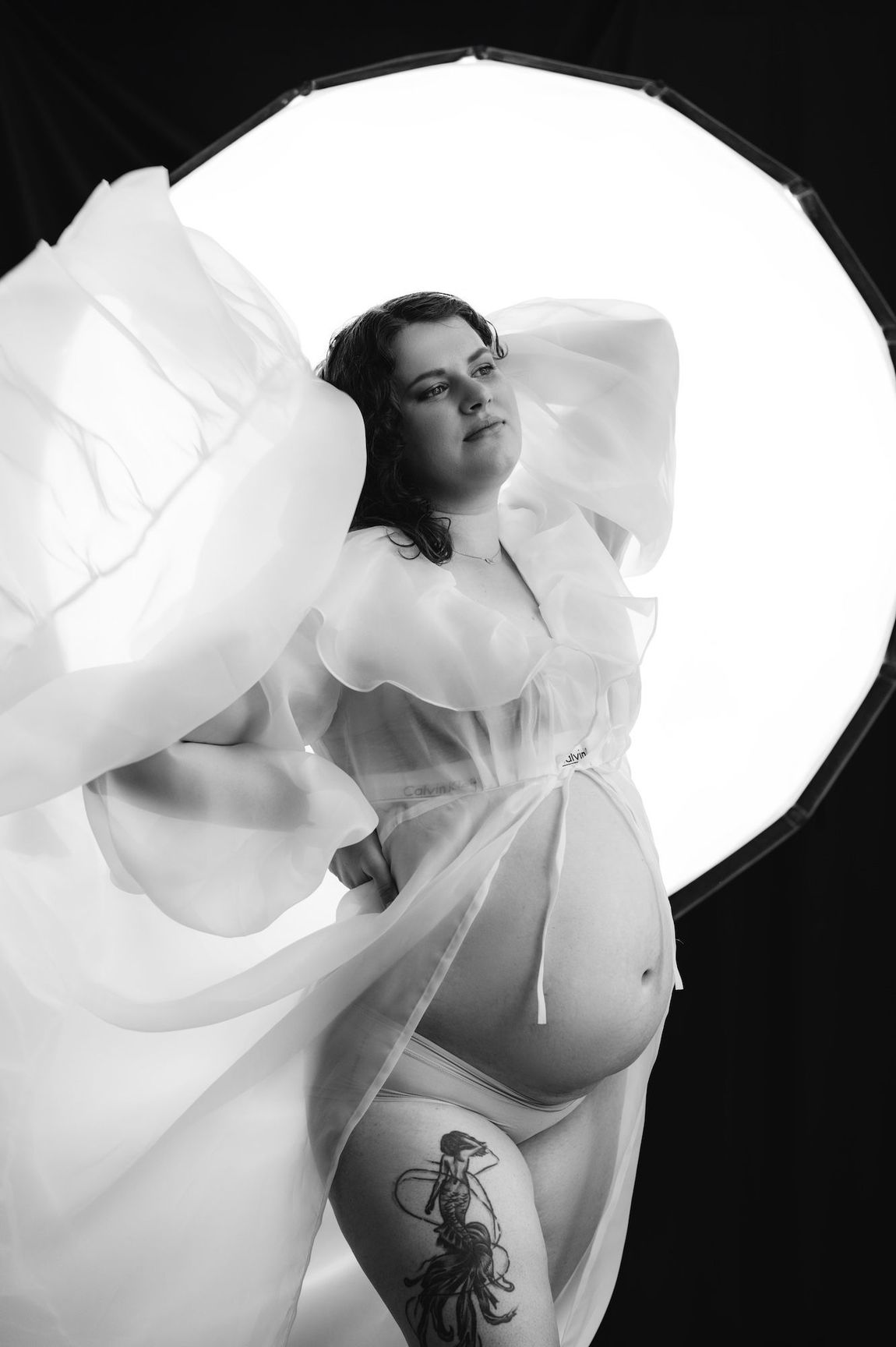 A black and white photo of a pregnant woman wearing a flowing white dress, capturing a moment of serene beauty.