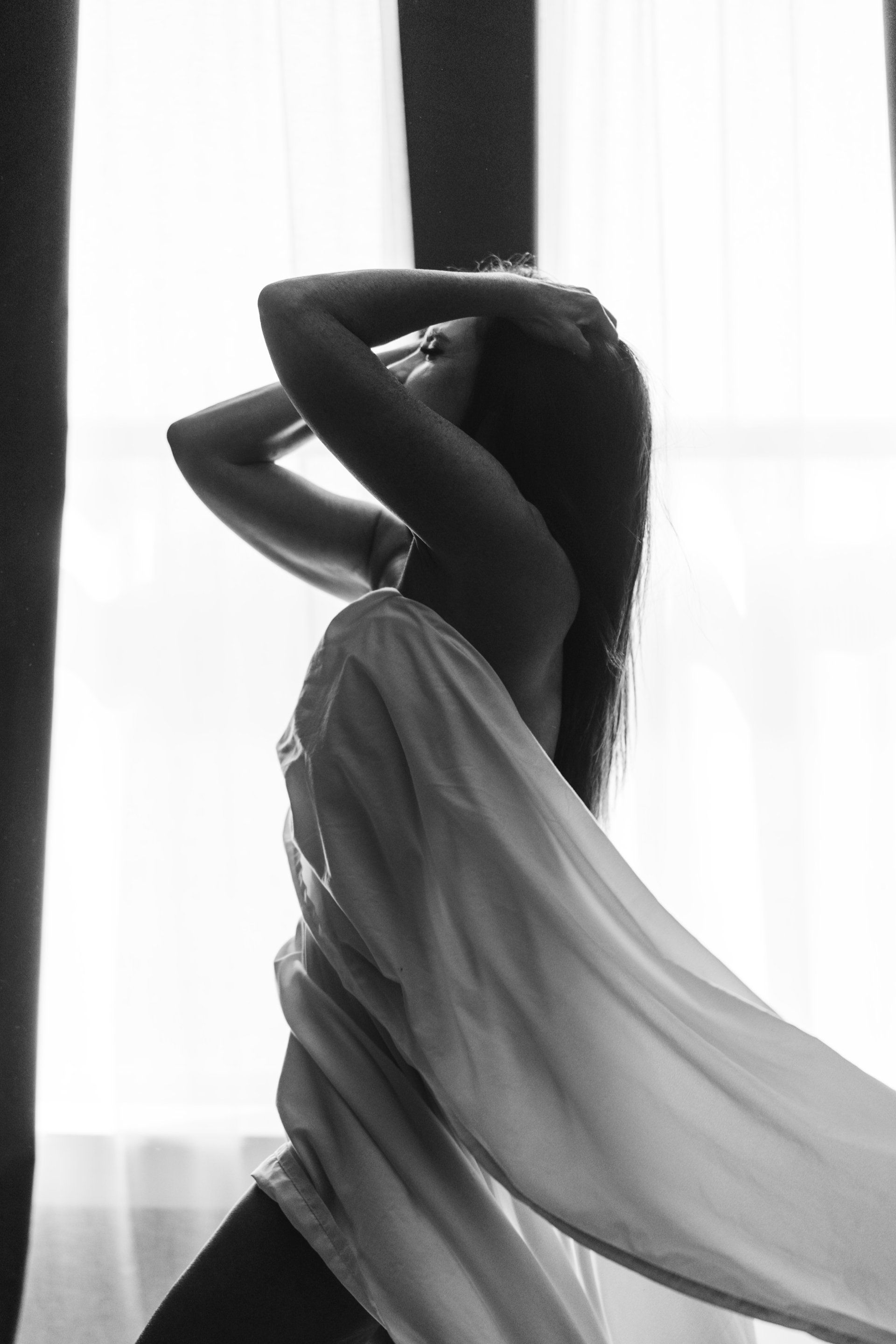 woman standing in front of window with hands in hair and sheet billowing behind her