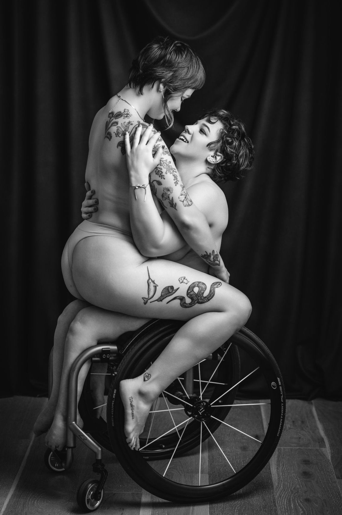 Two women seated in a wheelchair, both without clothing, in a serene and intimate setting.