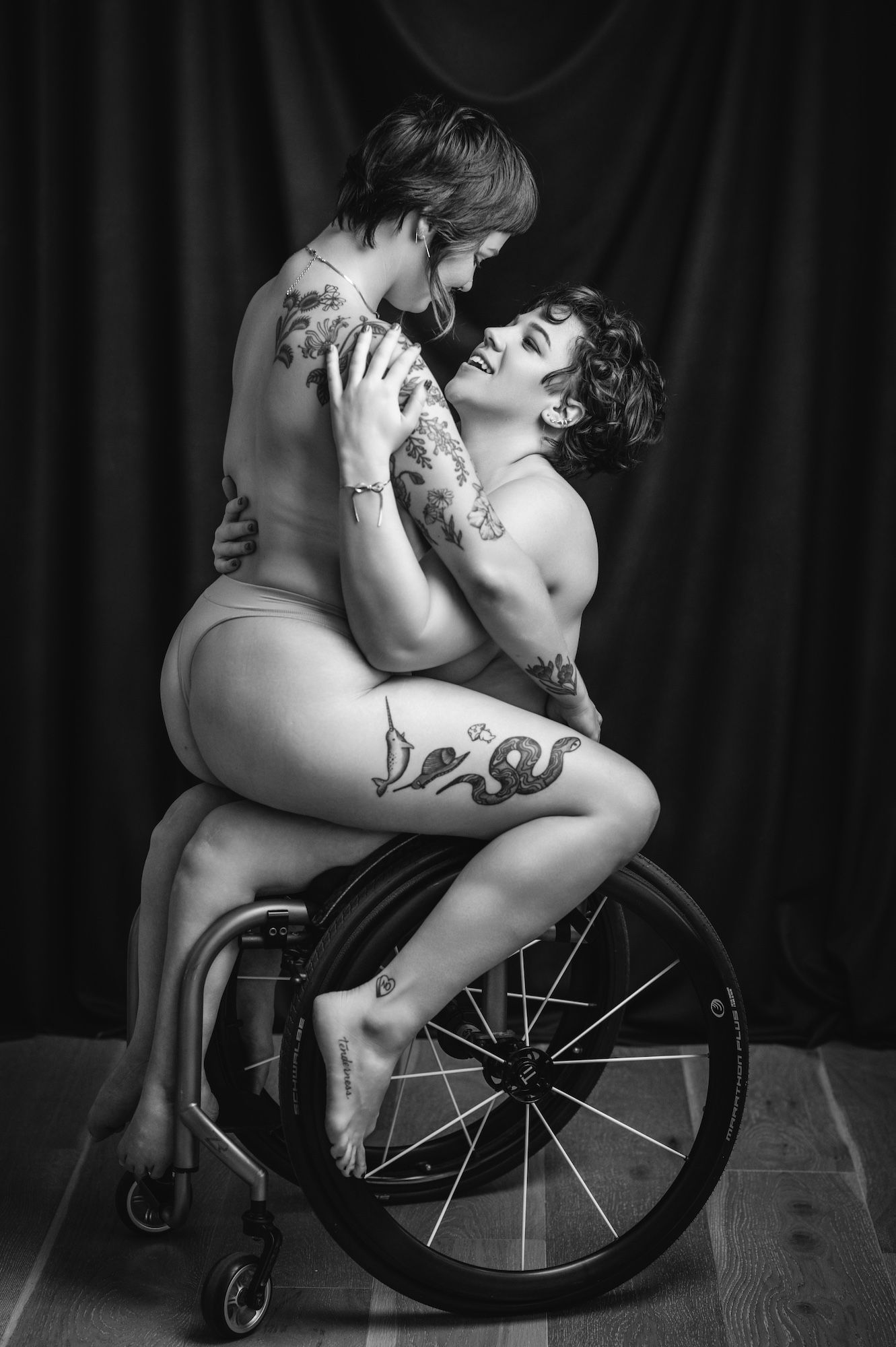 Two women seated in a wheelchair, both without clothing, in a serene and intimate setting.