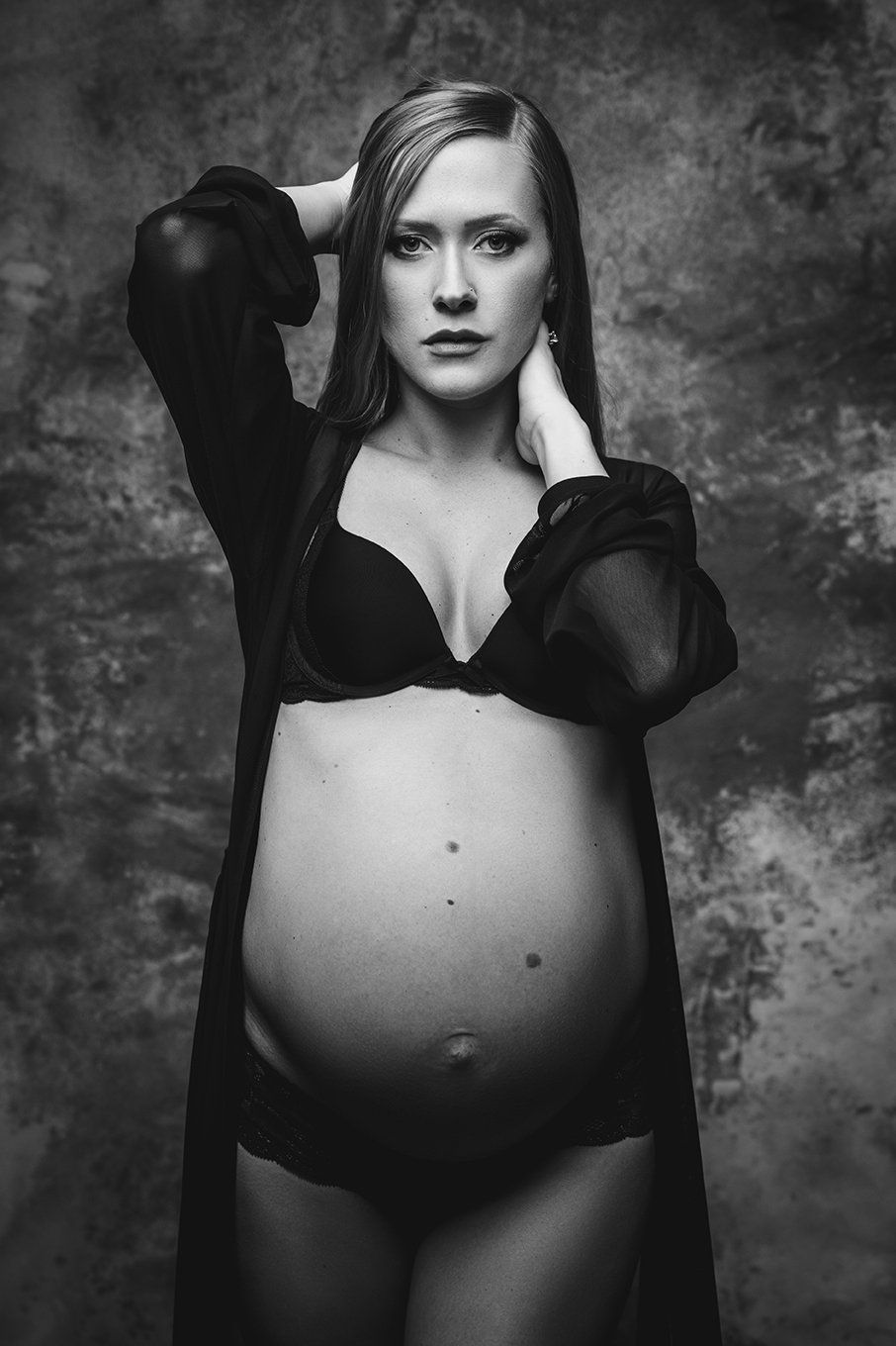 maternity photo of woman standing in front of backdrop, looking into camera with hand on neck