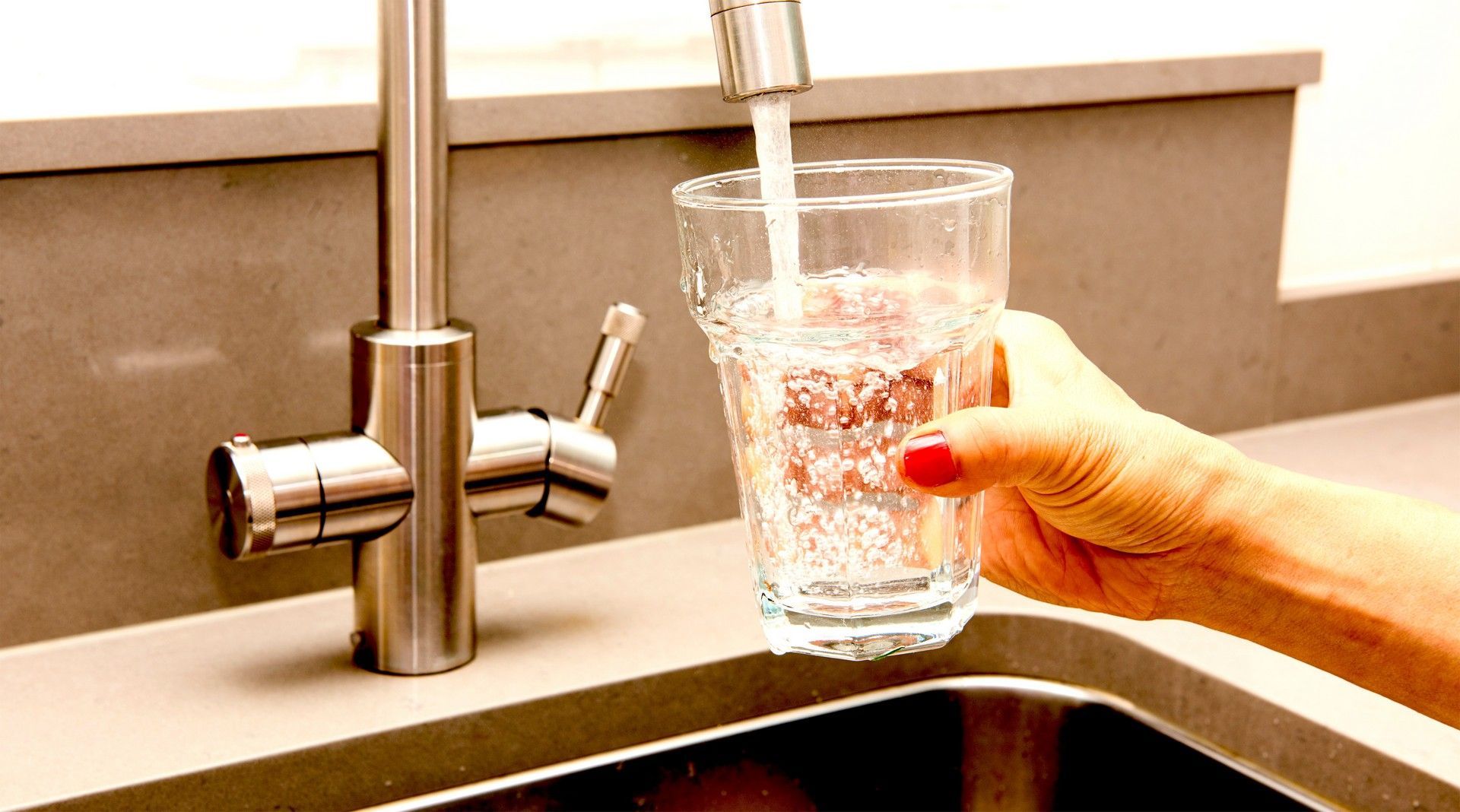 Persona che riempie un bicchiere trasparente con l'acqua di un rubinetto argentato in cucina.