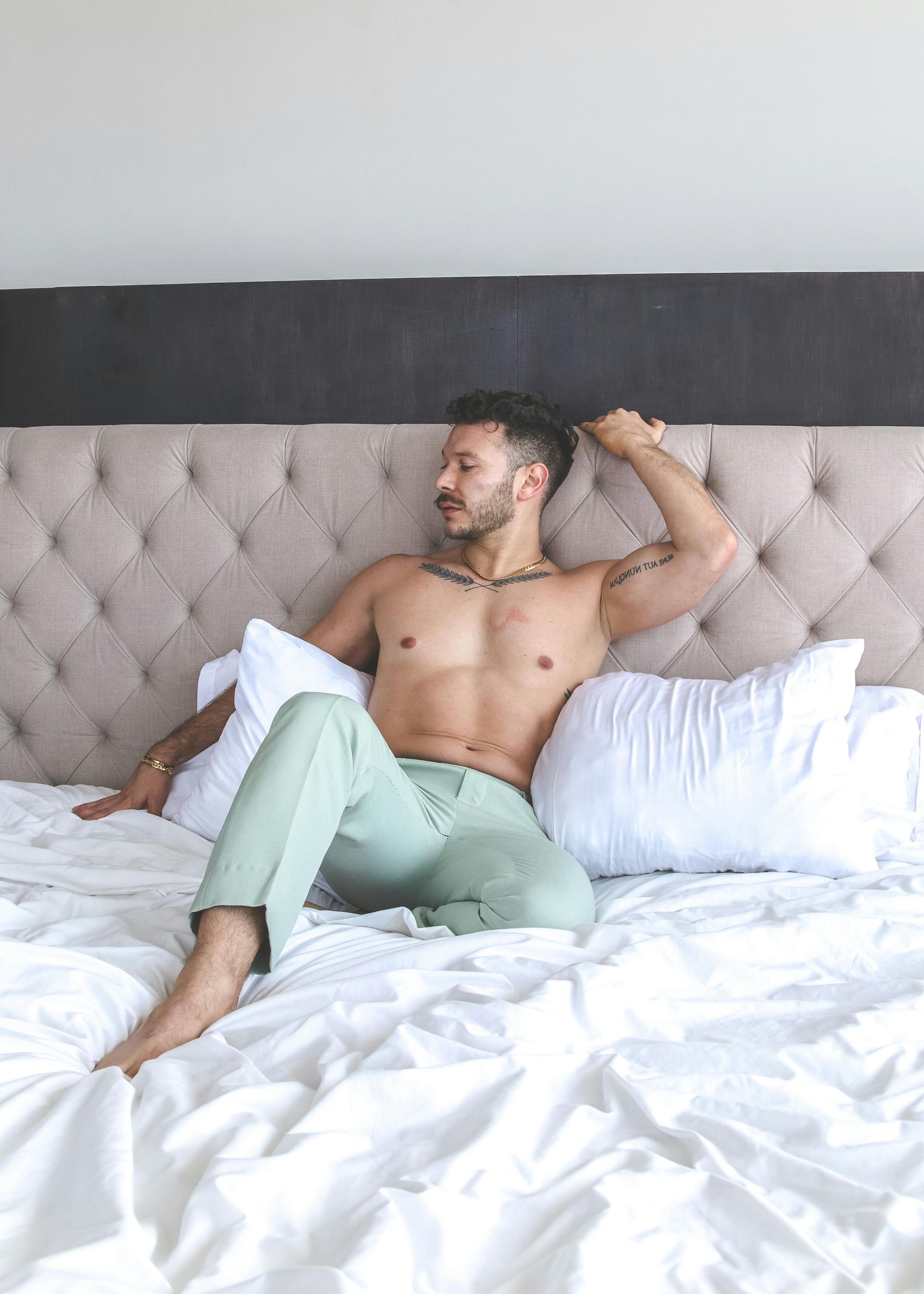 A shirtless man is sitting on a bed with white sheets.