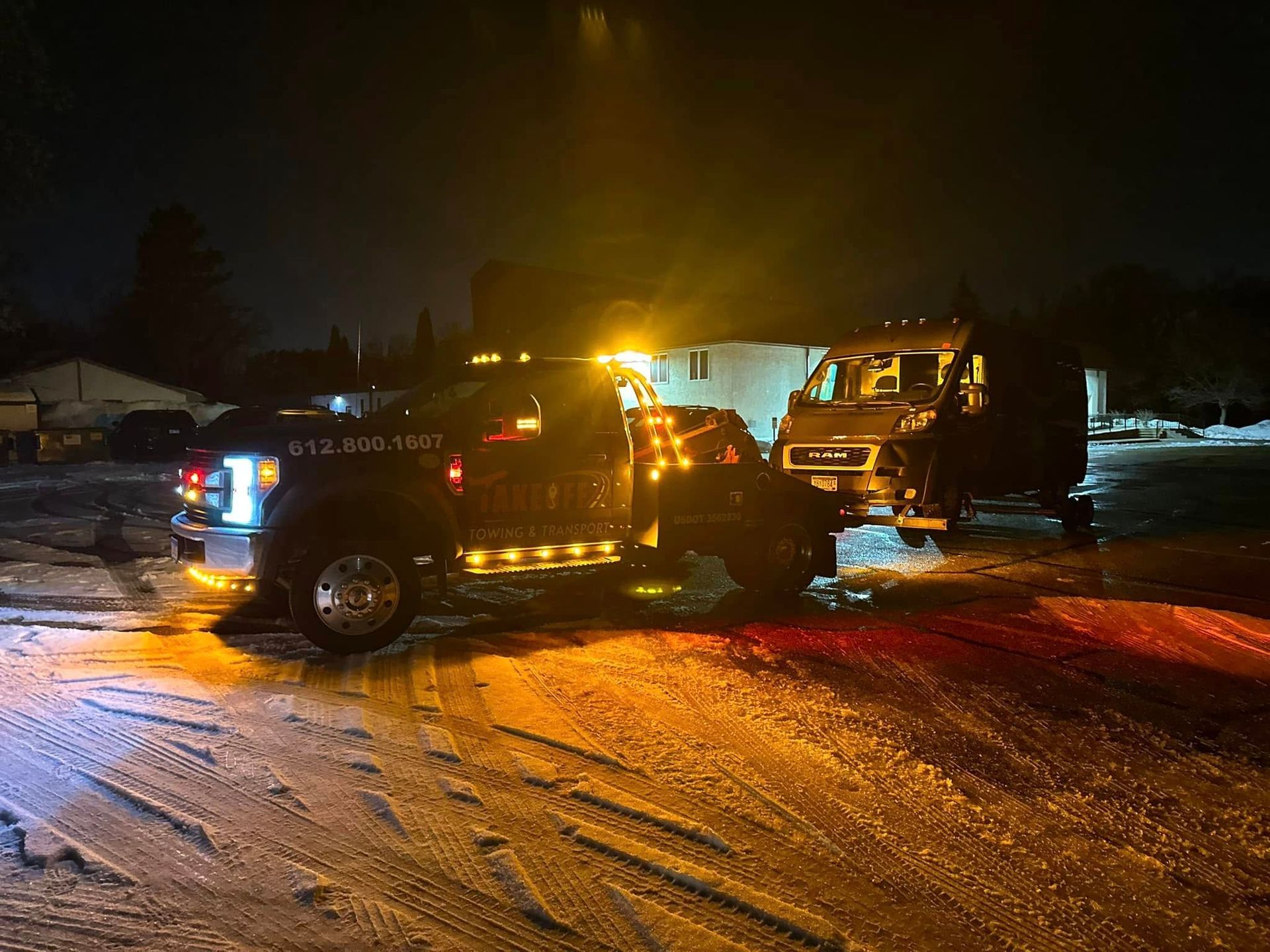 Tow truck hauling a vehicle at night. Snow-covered ground, emergency lights illuminated, dark setting.
