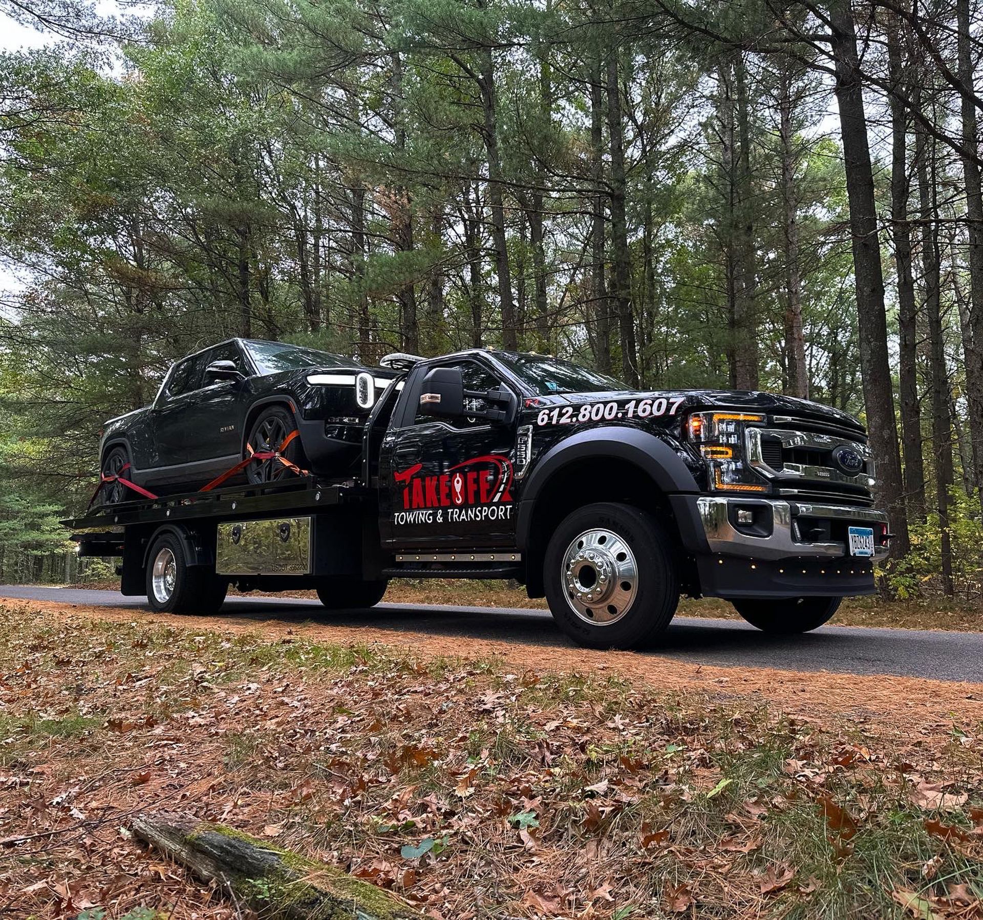 Tow truck with a black car loaded, driving on a road in a forest.