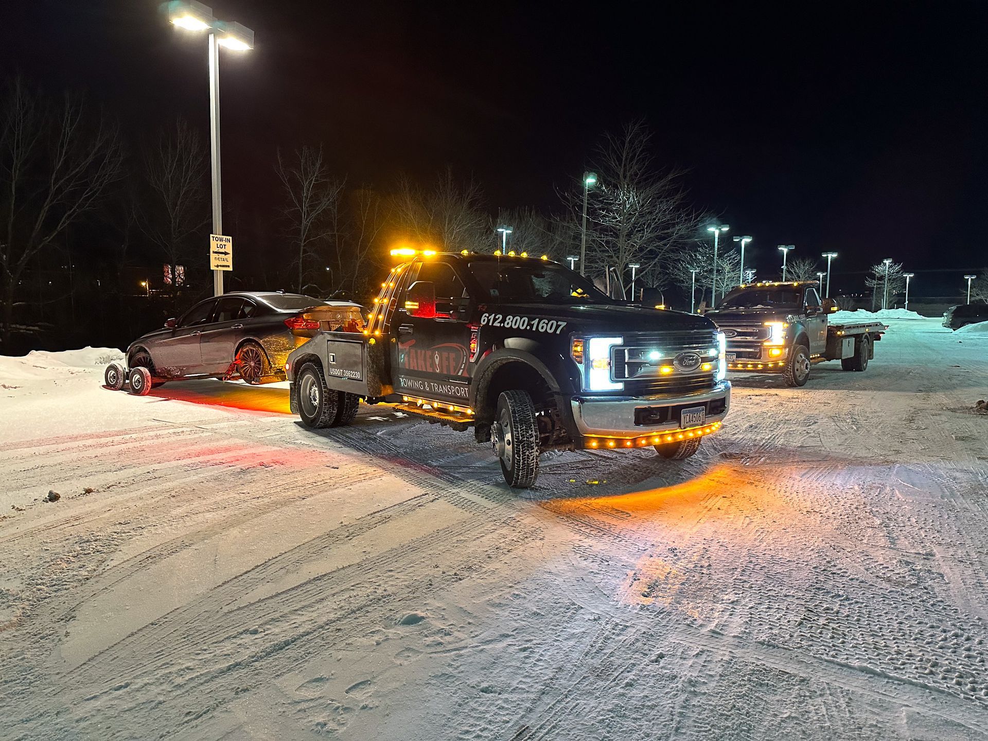 Tow truck at night towing a car in a snowy parking lot with flashing amber lights.