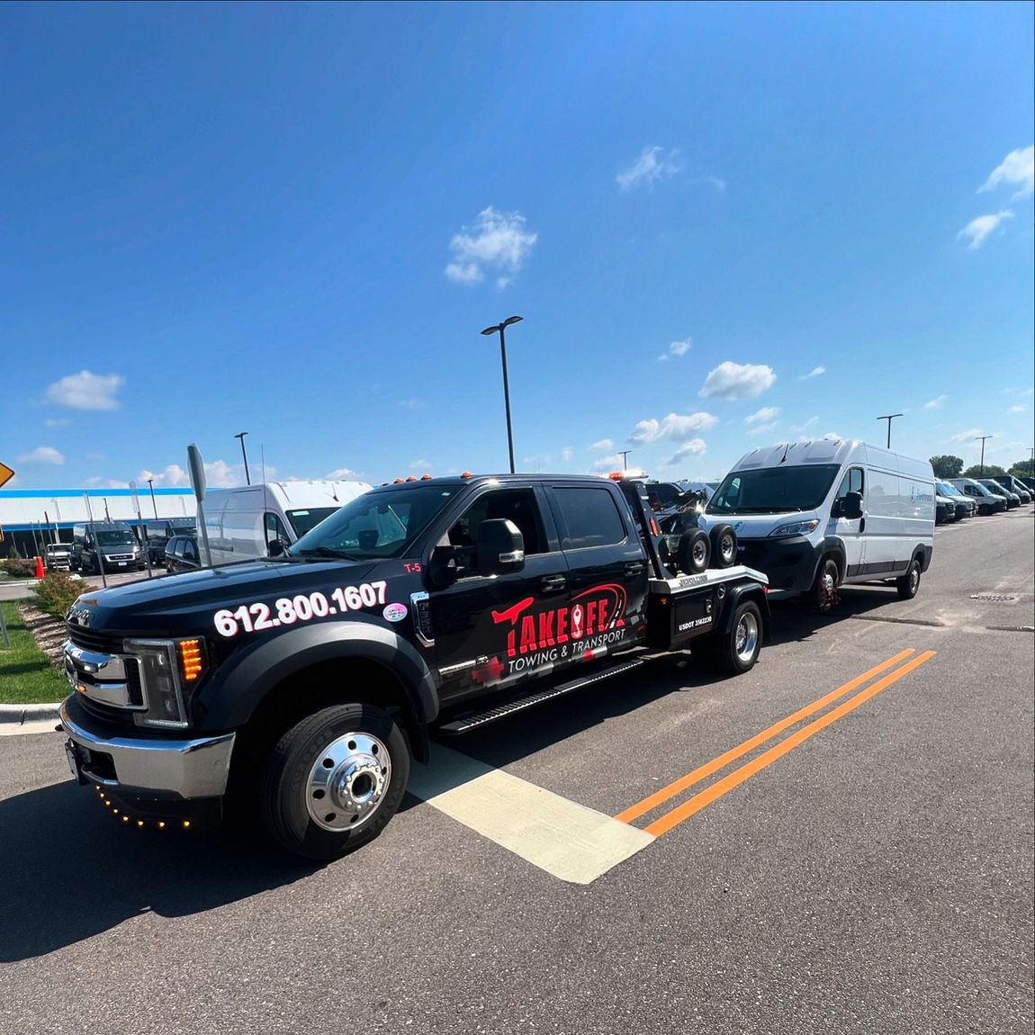 Black tow truck with a white van in tow on a sunny day. The tow truck has the phone number 612.800.1607 on it.