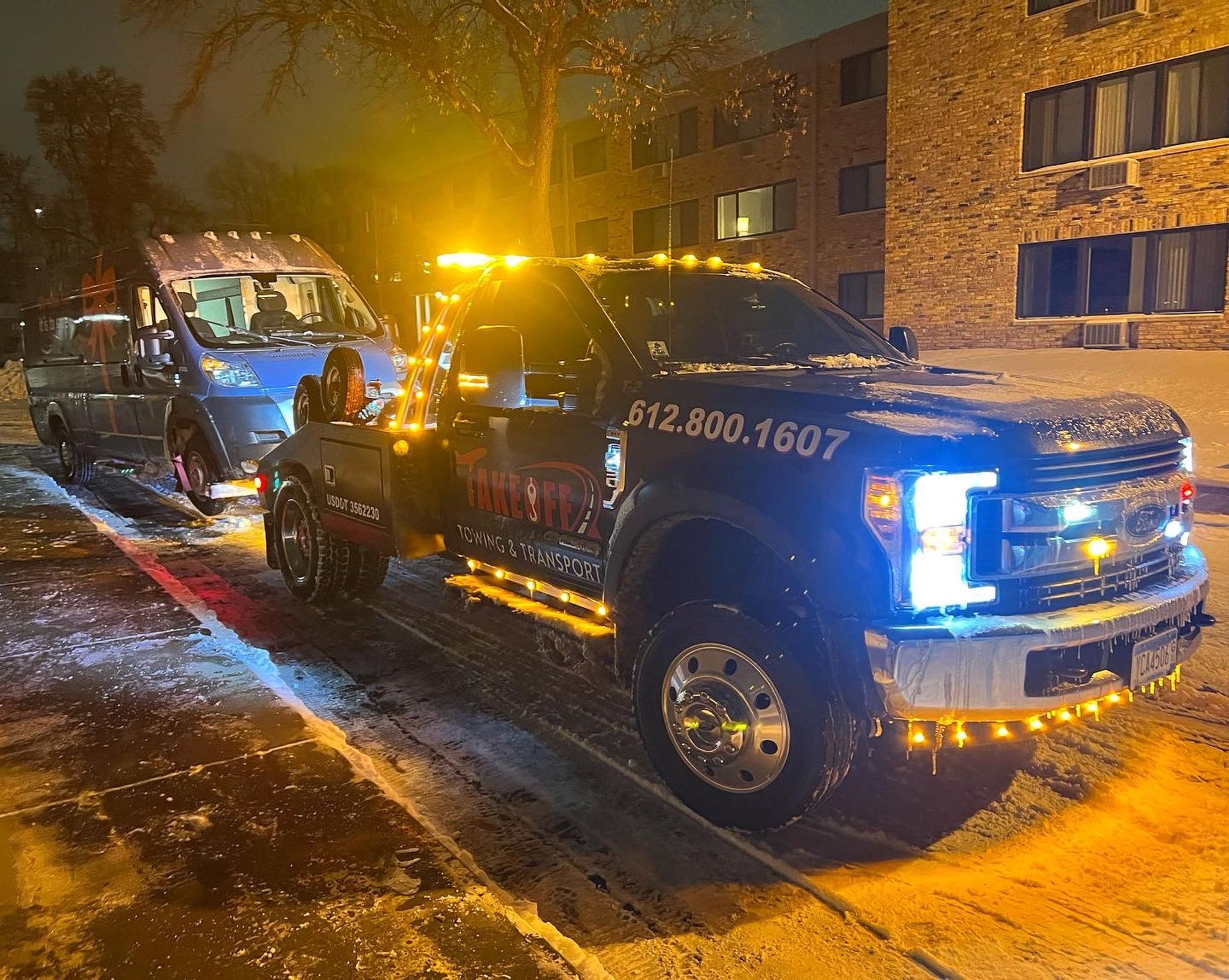 A tow truck towing a van at night in a snowy urban environment with flashing amber lights.