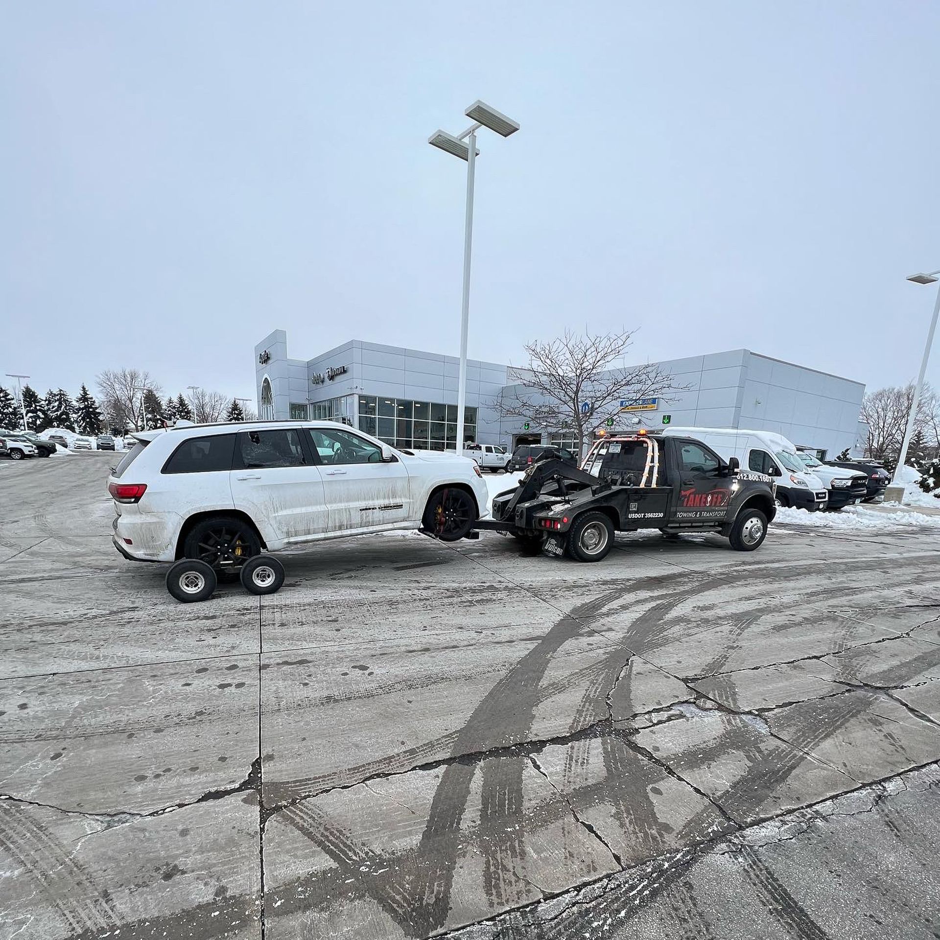 White SUV being towed by a tow truck in snowy parking lot, dealership in the background.