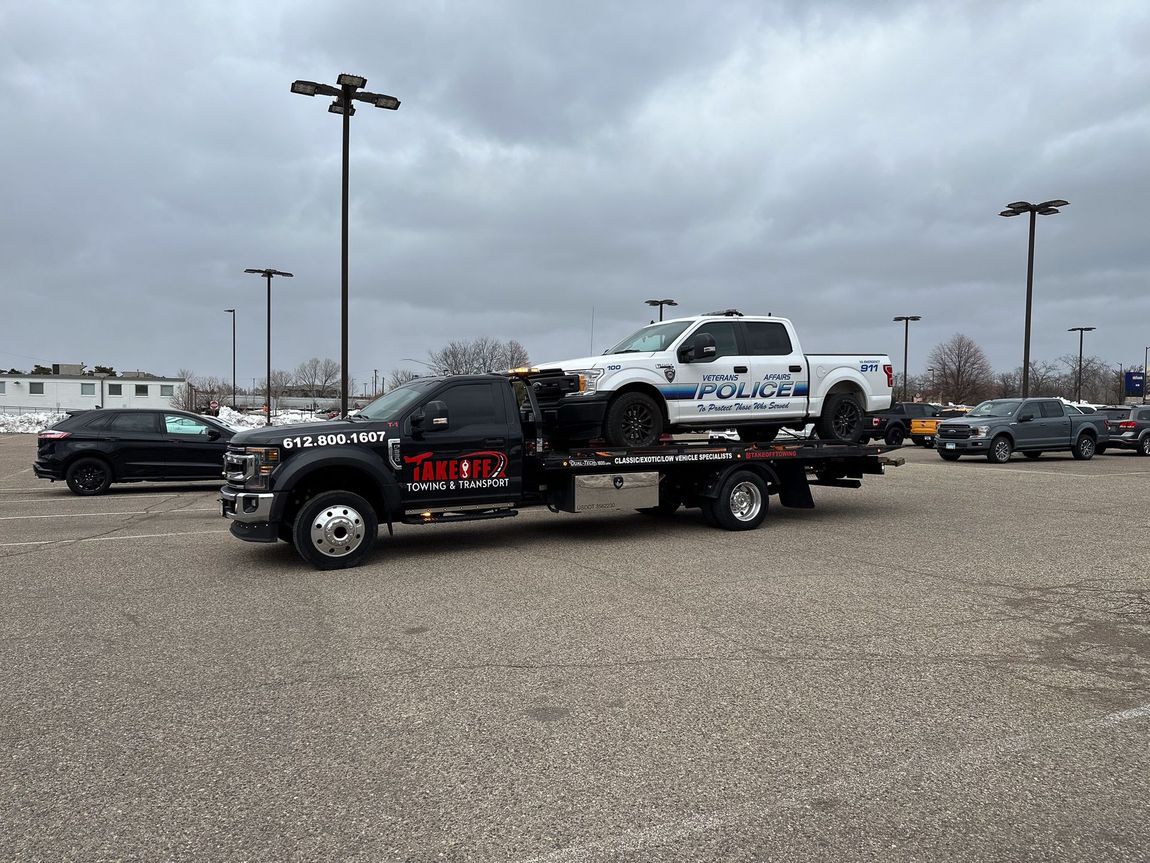 Tow truck carrying a white police pickup truck in a parking lot under a cloudy sky.