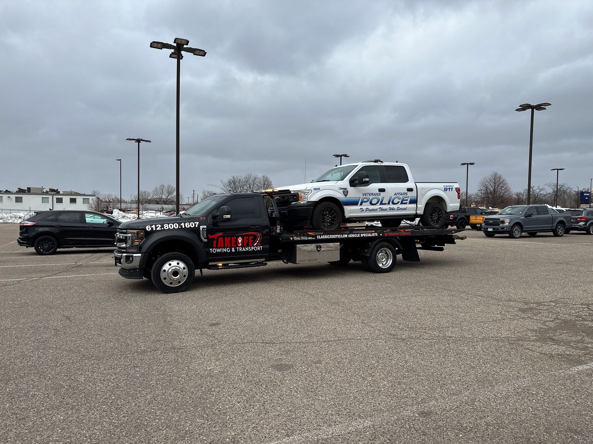 Tow truck carrying a white police pickup truck in a parking lot on a cloudy day.
