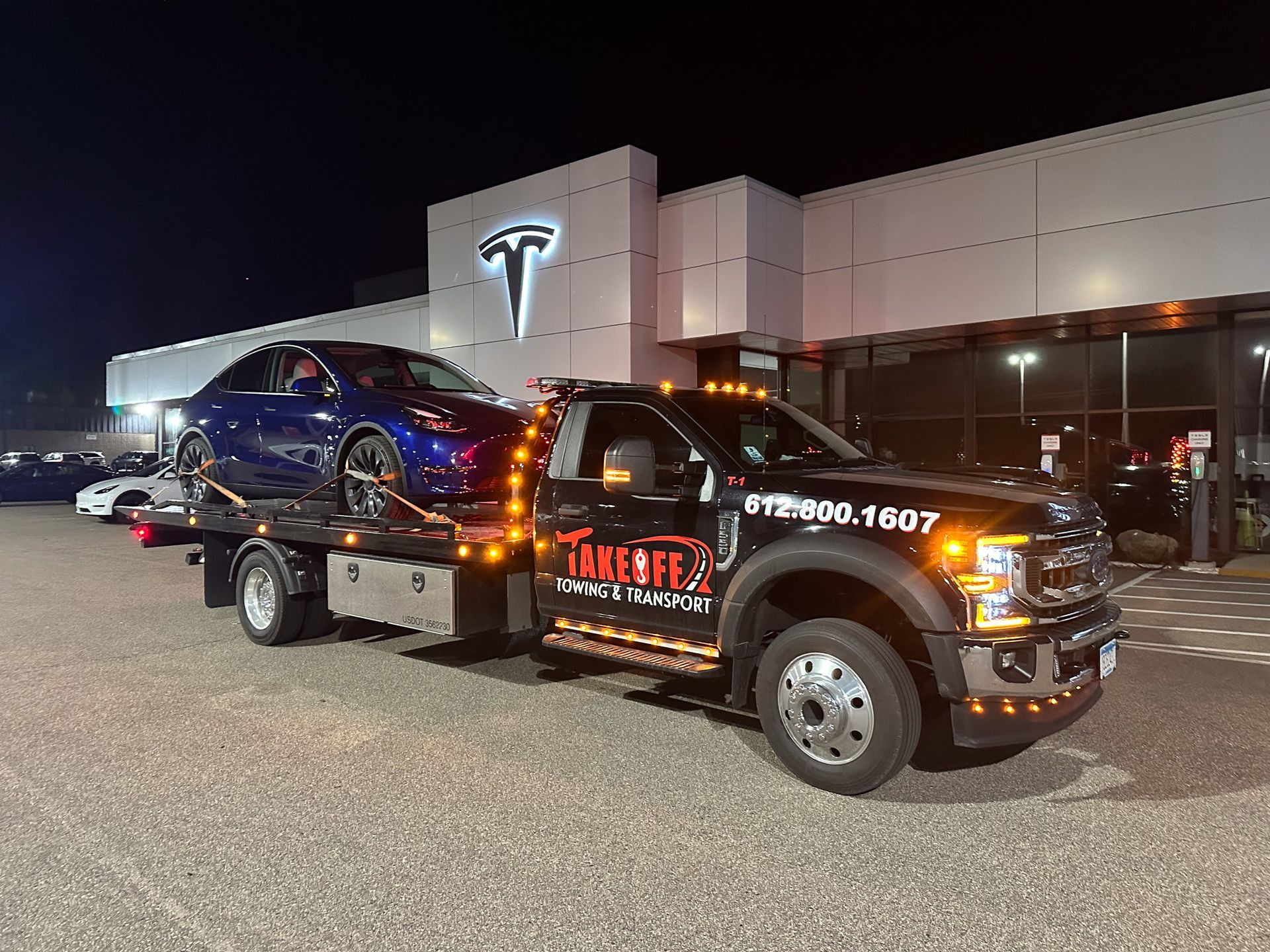 Tow truck with a blue Tesla at a Tesla dealership at night.