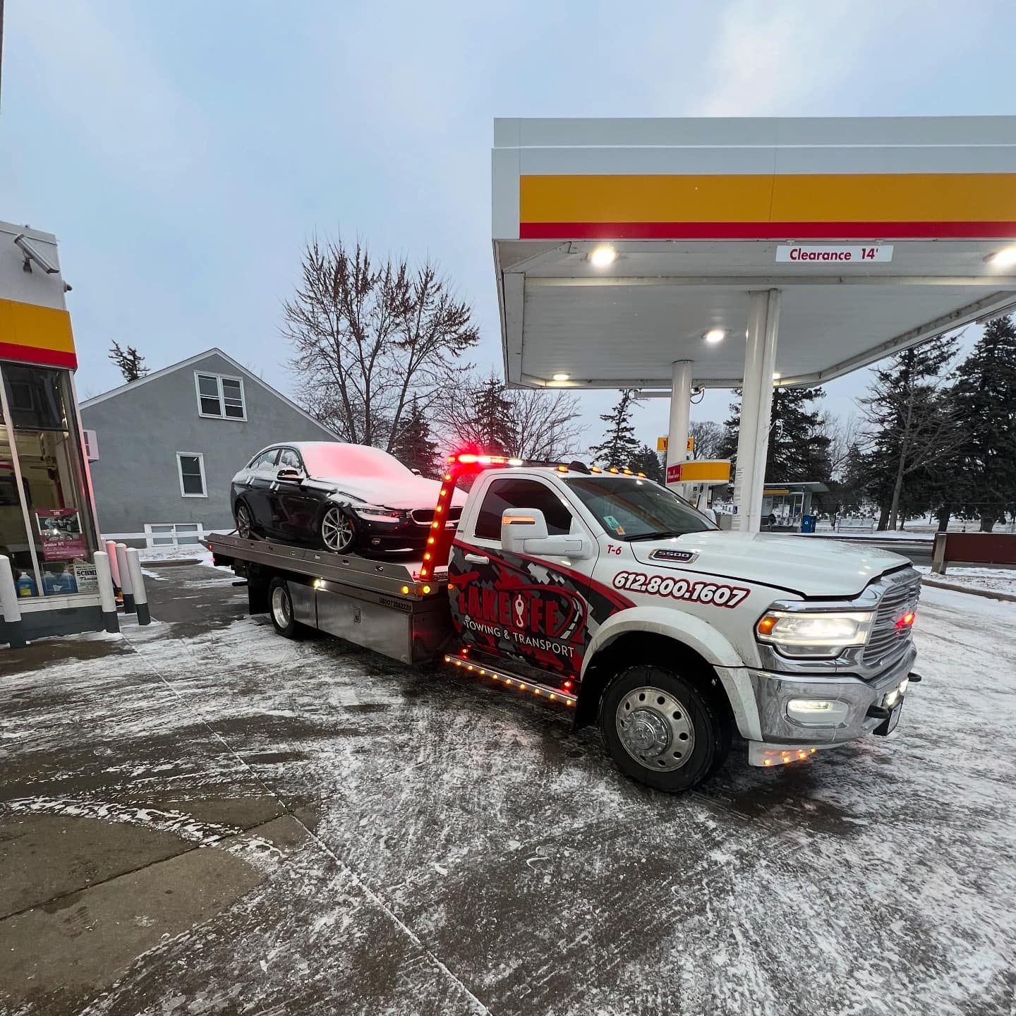 Tow truck with a car, at a Shell gas station in snowy weather. The truck's lights are on.