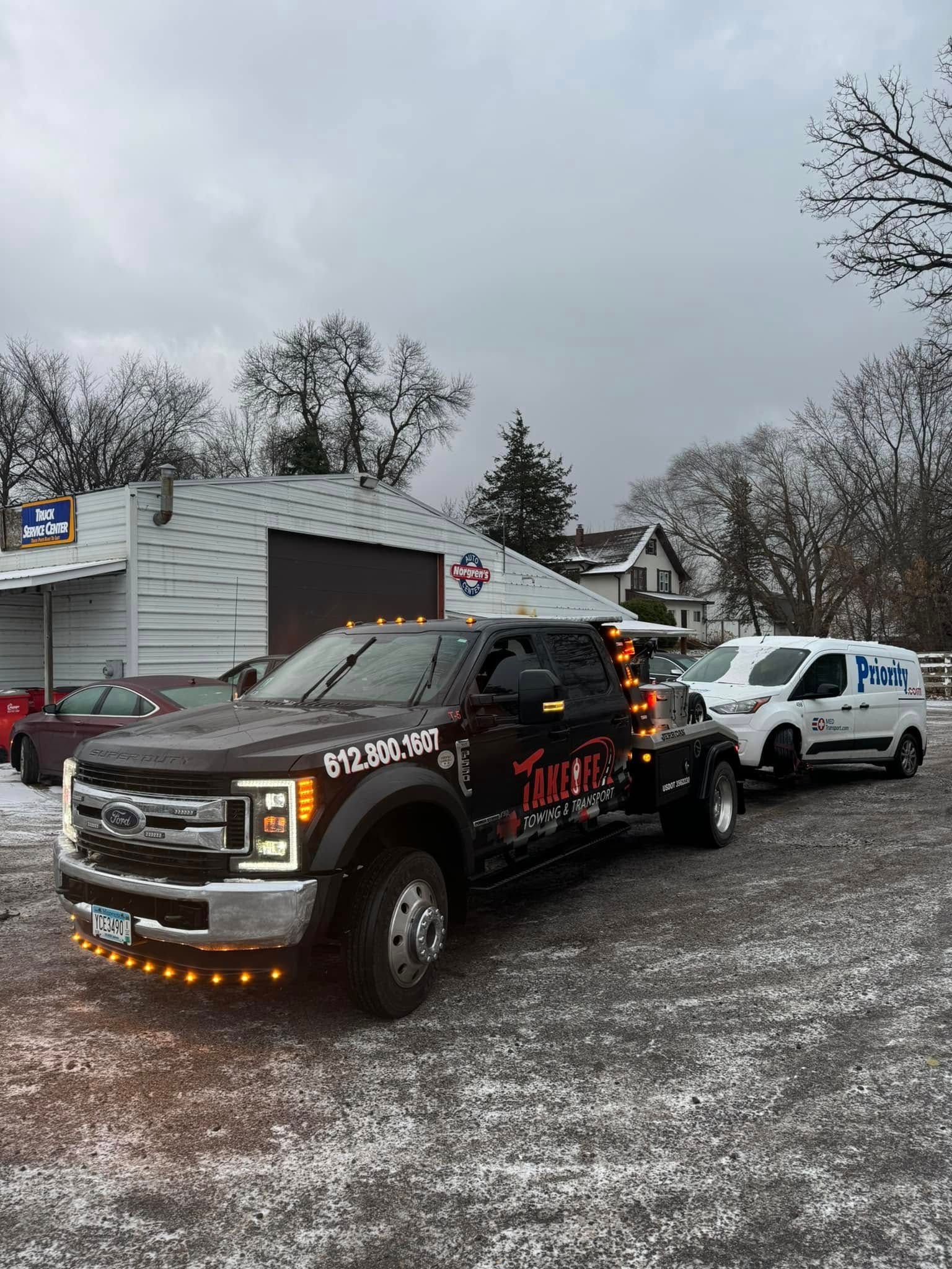 Tow truck towing a white van on a snow-covered lot in front of a building under an overcast sky.