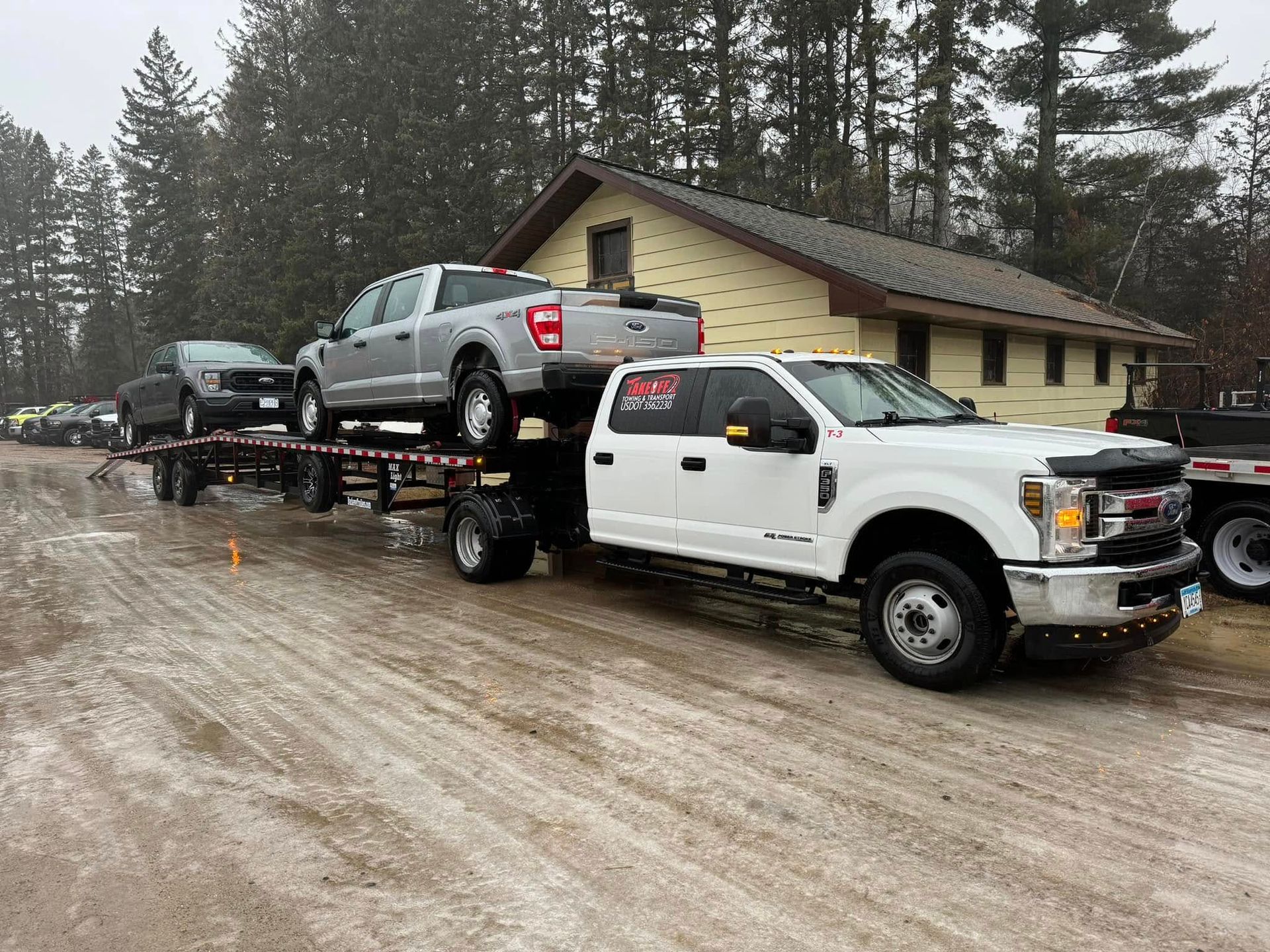 White truck towing a trailer loaded with two silver trucks, parked on a snowy road near a building.