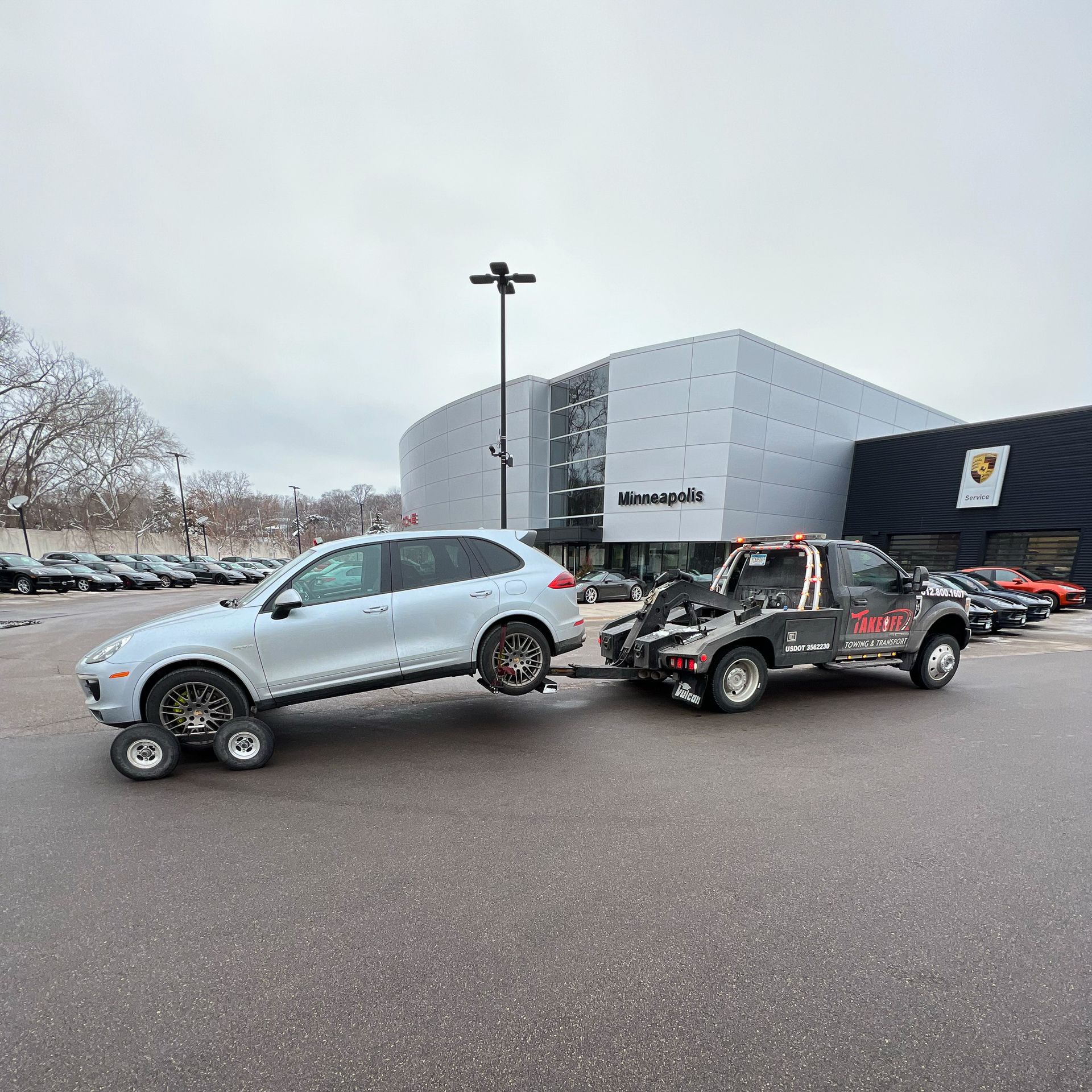 A light silver SUV being towed by a black tow truck in front of a Porsche dealership on a cloudy day.