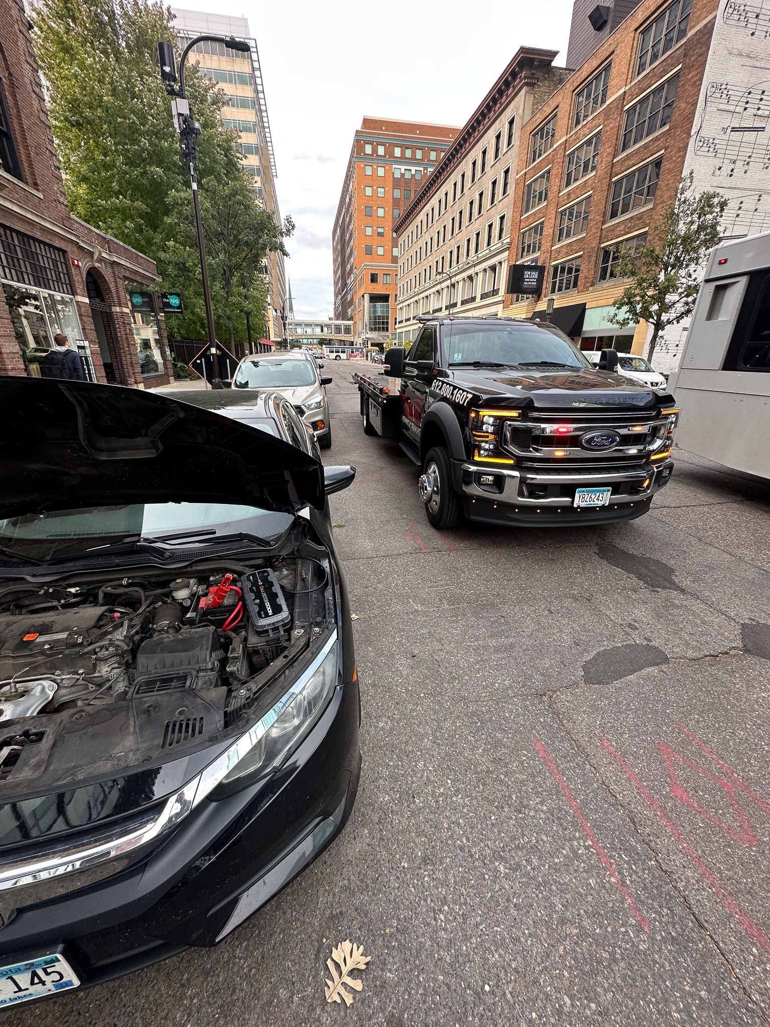 Black car with open hood, city street with truck, buildings in the background.