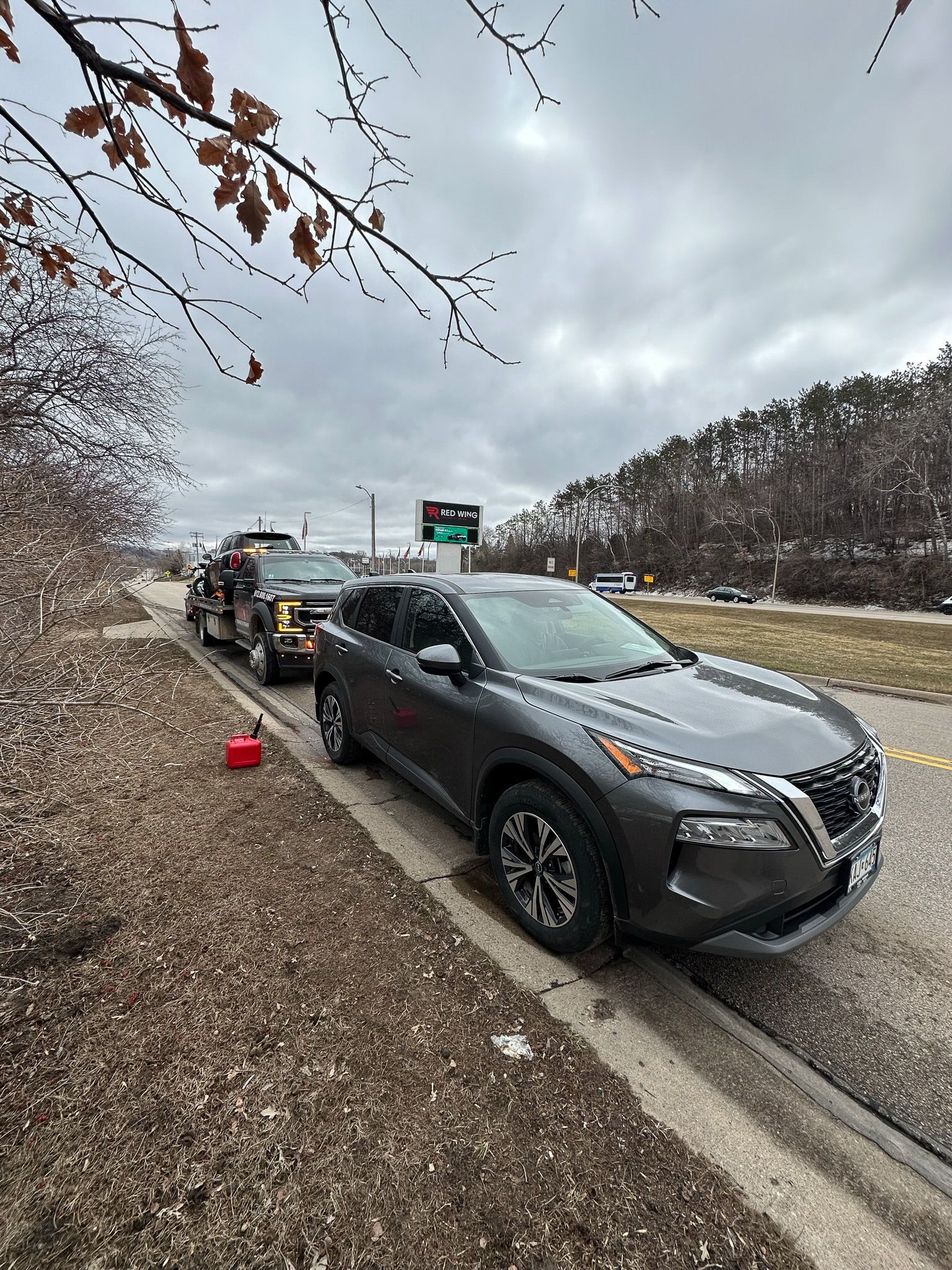 Gray Nissan Rogue parked on roadside, being towed. Fuel can on ground. Overcast day.