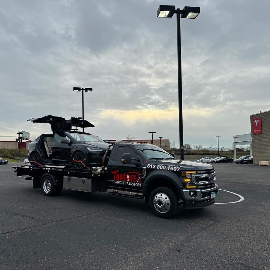 Black Tesla SUV on a tow truck, doors open, parked outside a Tesla dealership on a cloudy day.