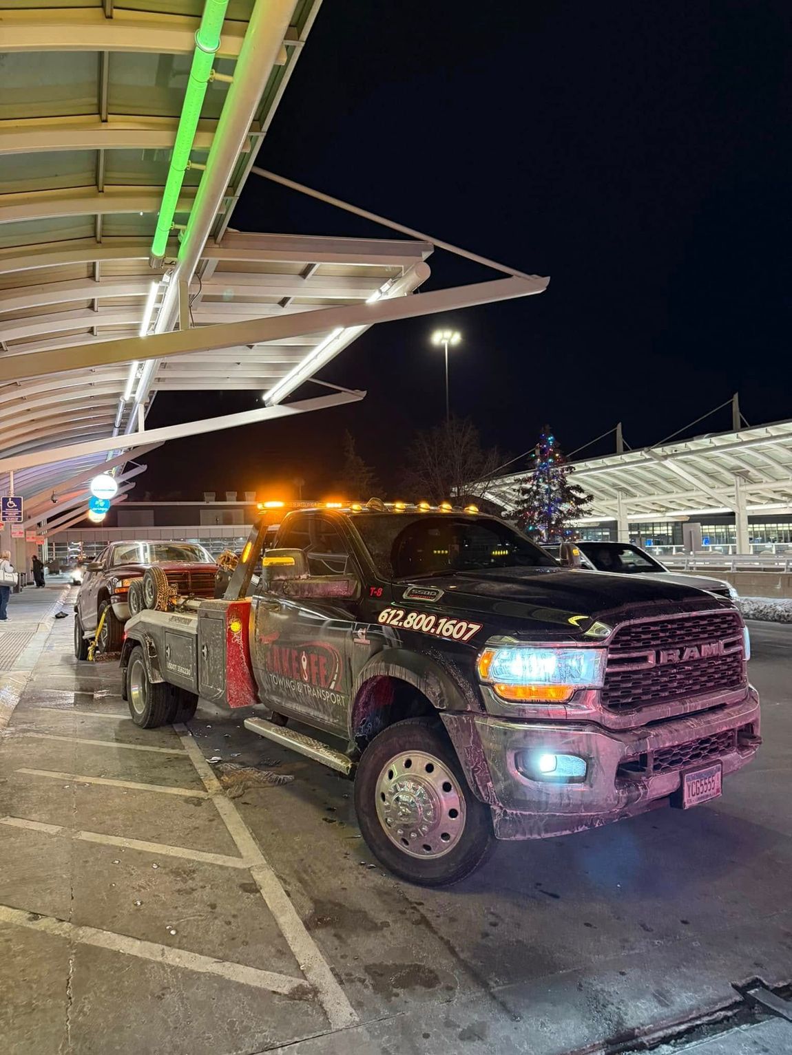 Tow truck with flashing lights towing a car at night, parked under a covered area.