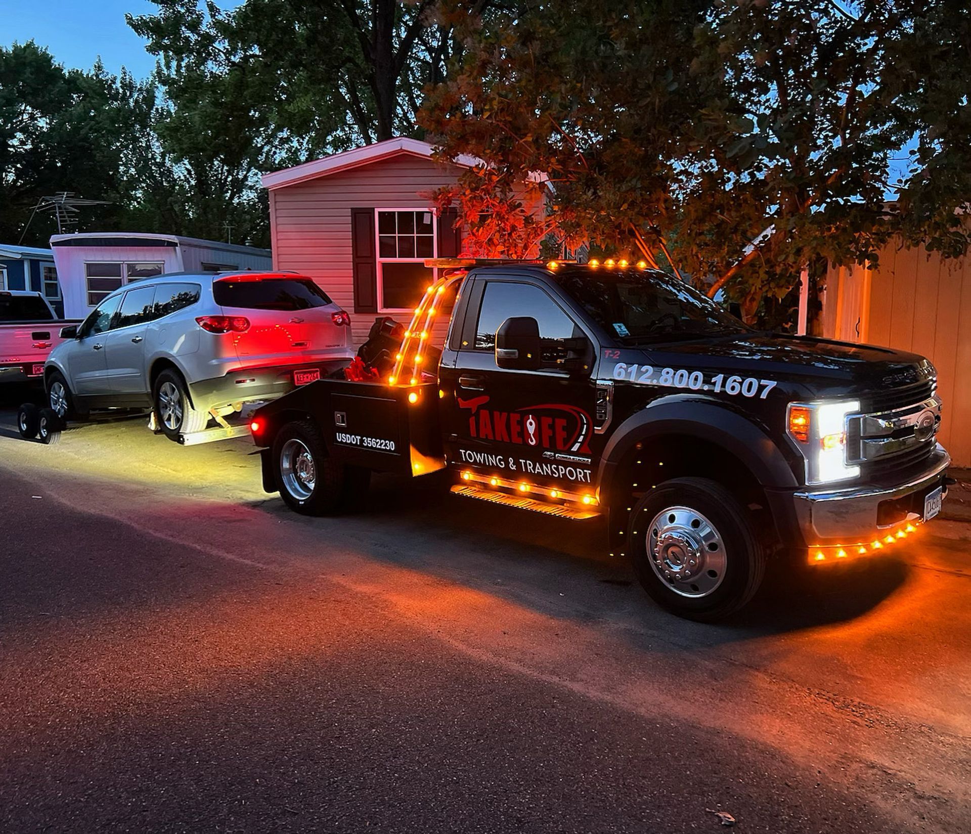 Black tow truck towing a white SUV on a residential street at dusk, illuminated by orange lights.