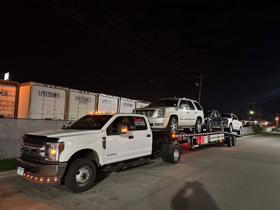 White truck towing three vehicles on a trailer at night, near containers.