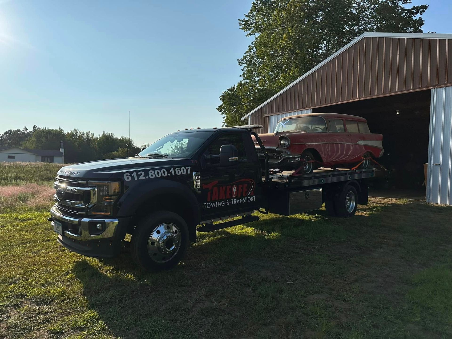 Black tow truck transports a vintage red station wagon beside a barn on a grassy field.