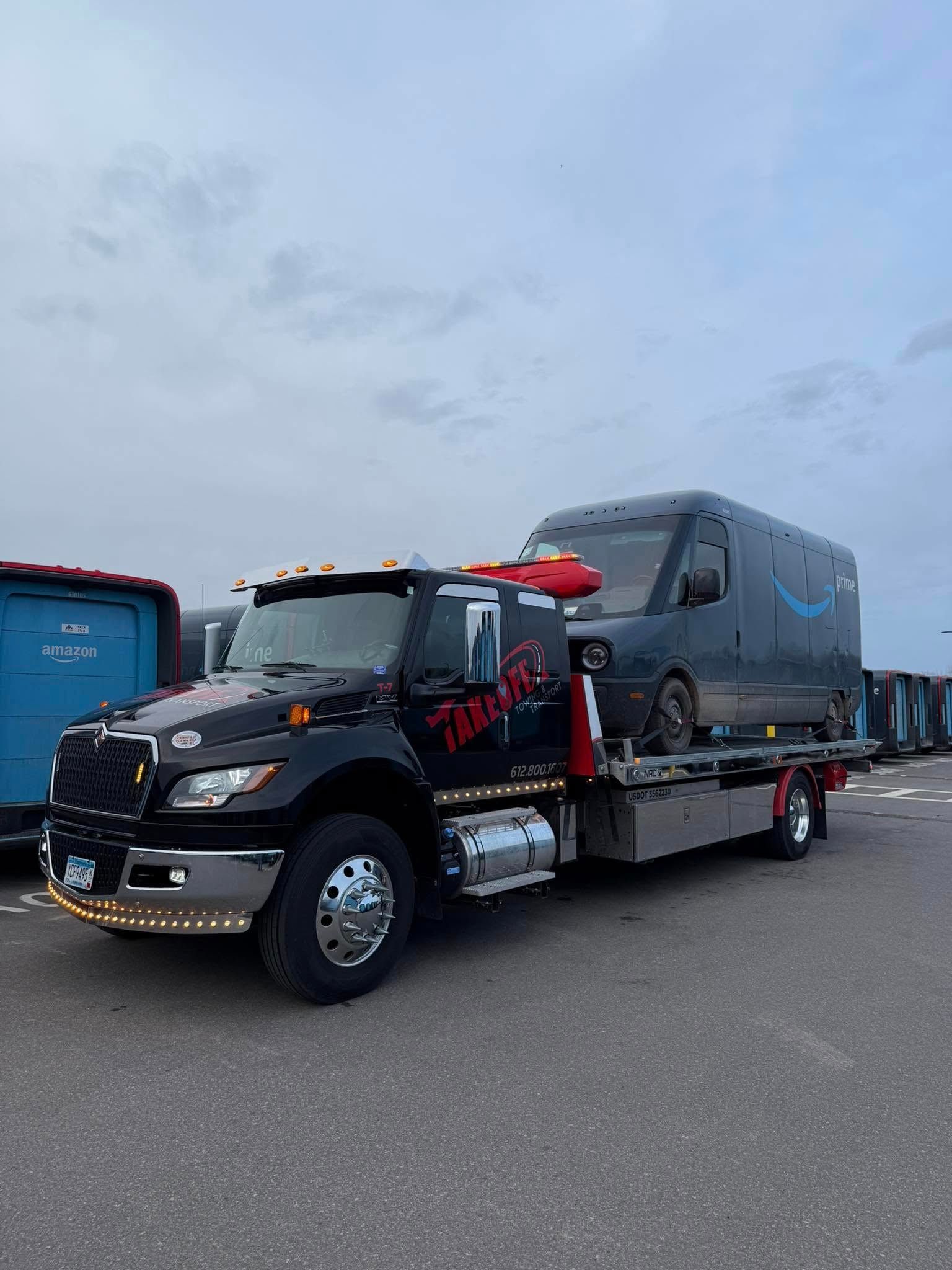 A black tow truck carrying a dark Amazon van.  Setting is a cloudy day in a parking lot.