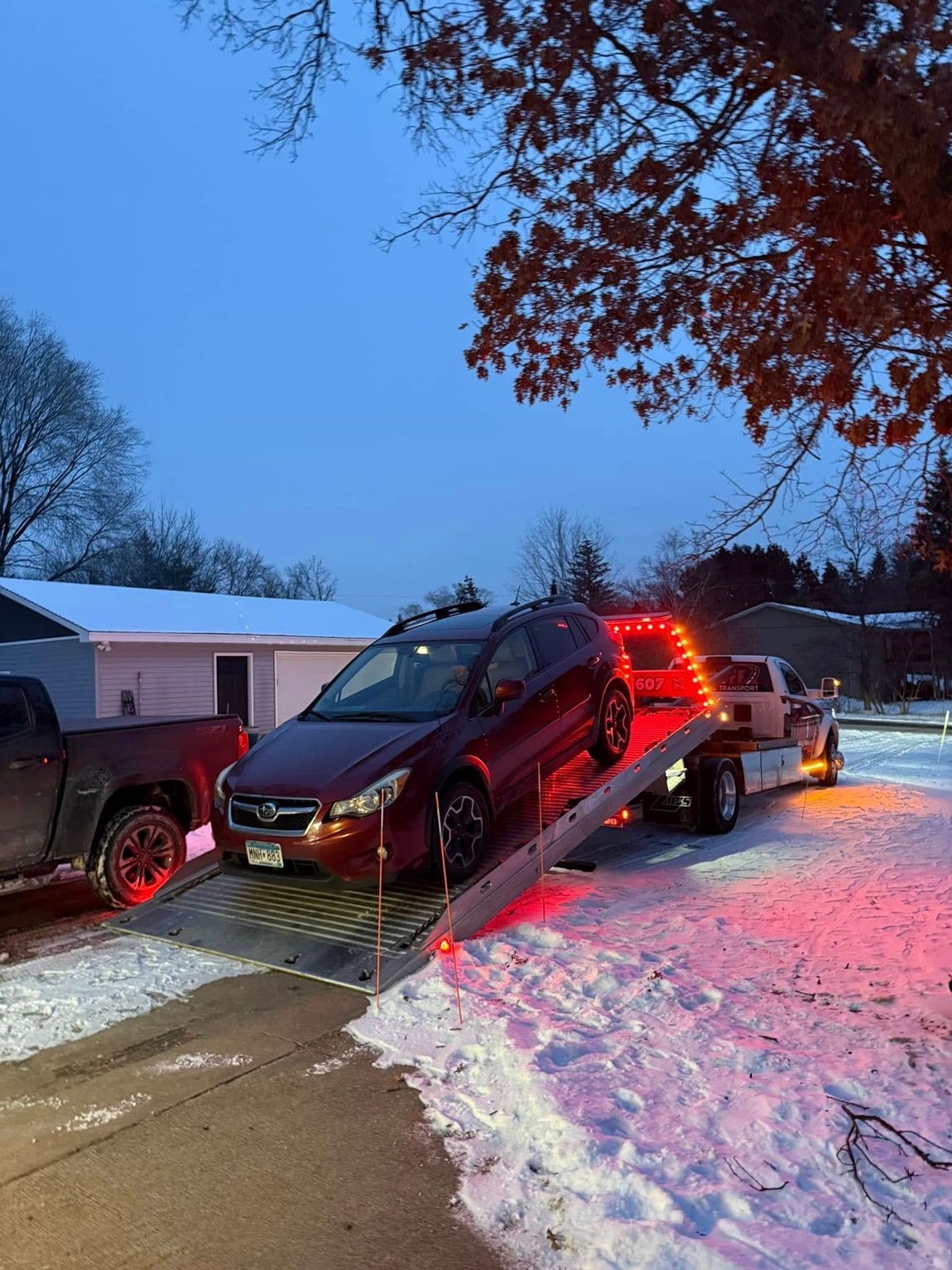 A maroon car being towed onto a flatbed truck on a snowy driveway at dusk.