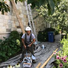 Un ouvrier du bâtiment, portant un casque blanc et une salopette, près d'un mur de pierre, d'une échelle et d'outils.